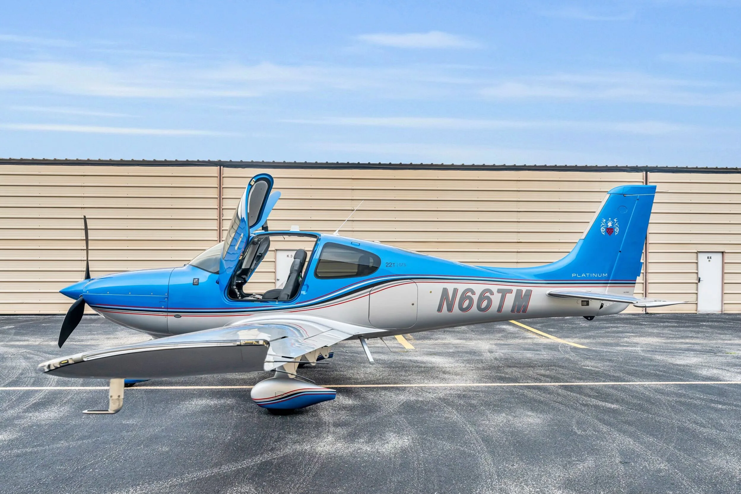 A blue and silver Cirrus SR22T GTS aircraft, registration N66TM, with its gull-wing doors open, parked on an asphalt ramp.