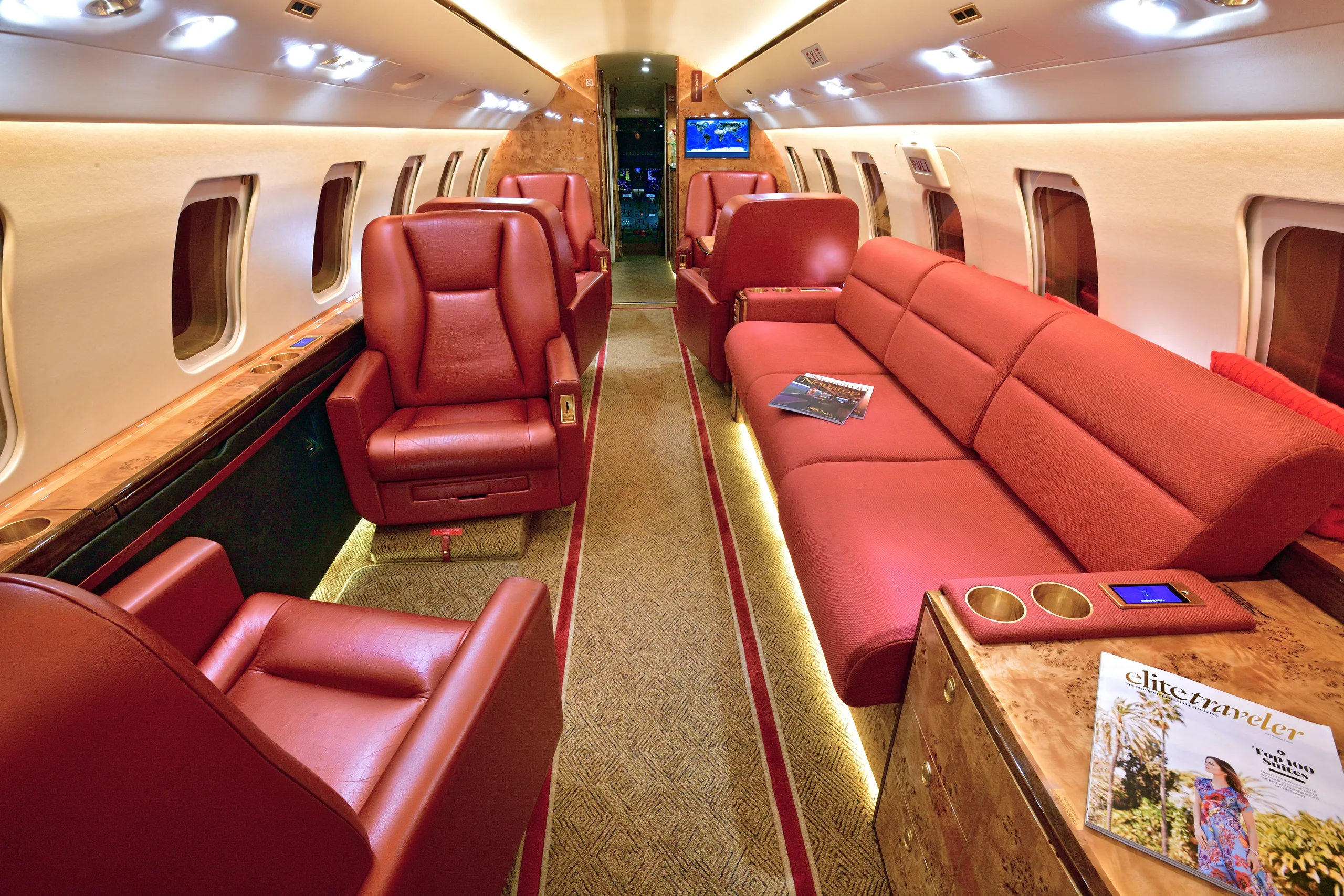 A wide view of a private jet's cabin interior with multiple red leather seats and a red sofa, illuminated by overhead lights.