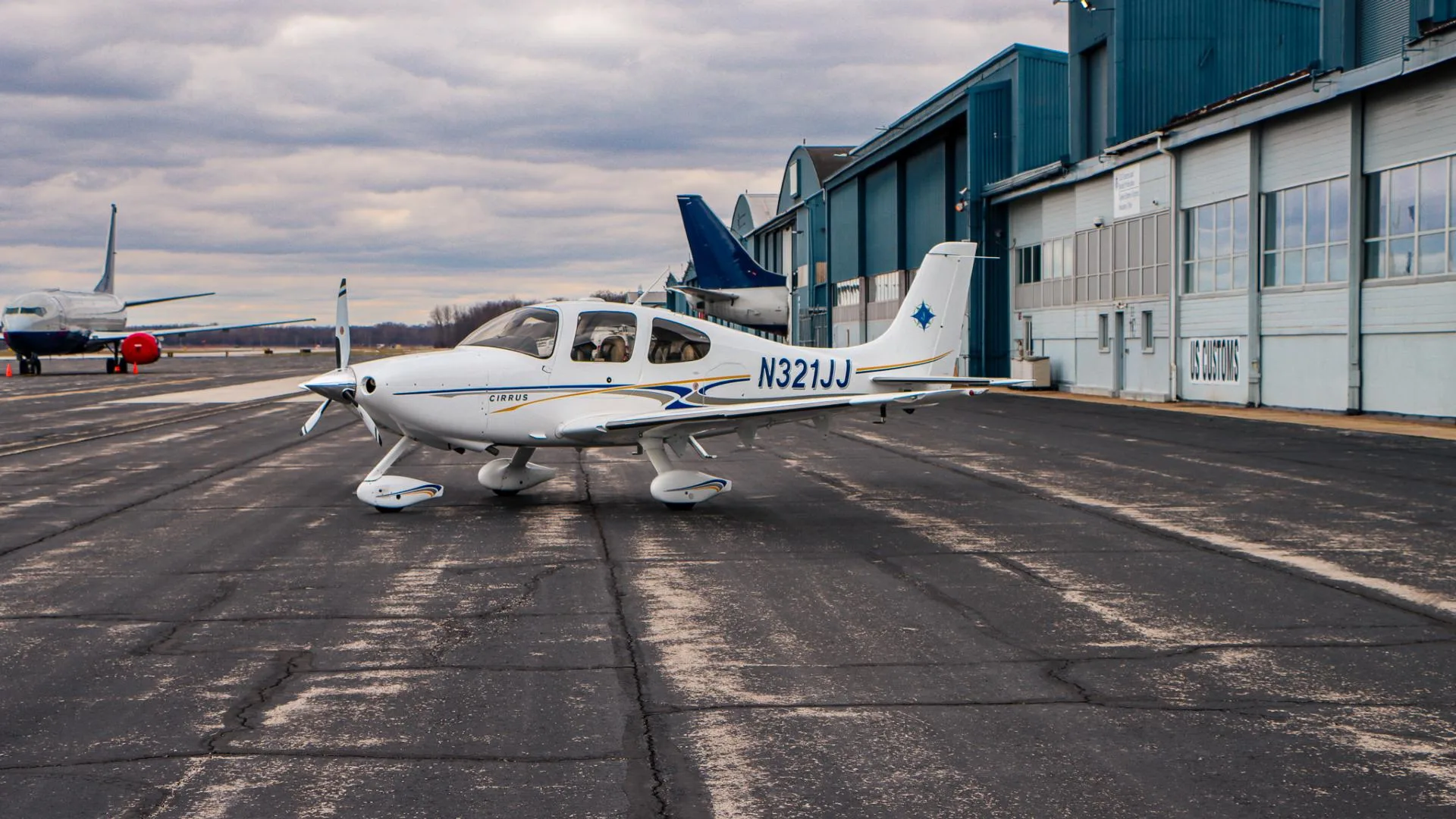 A white Cirrus aircraft, registration N321JJ, with blue and gold stripes, is parked on an airport tarmac.