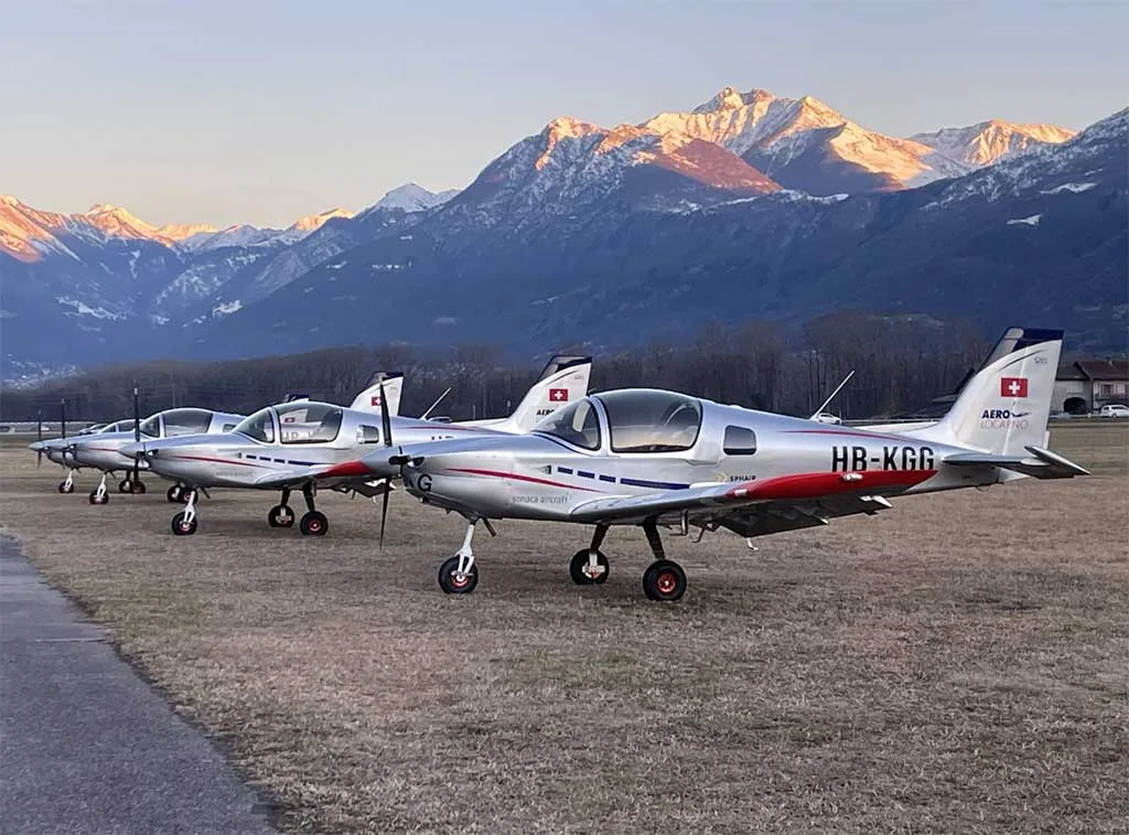 Three silver Sonaca 200 aircraft with red and blue accents parked on a grassy field with snow-capped mountains in the background at sunset.