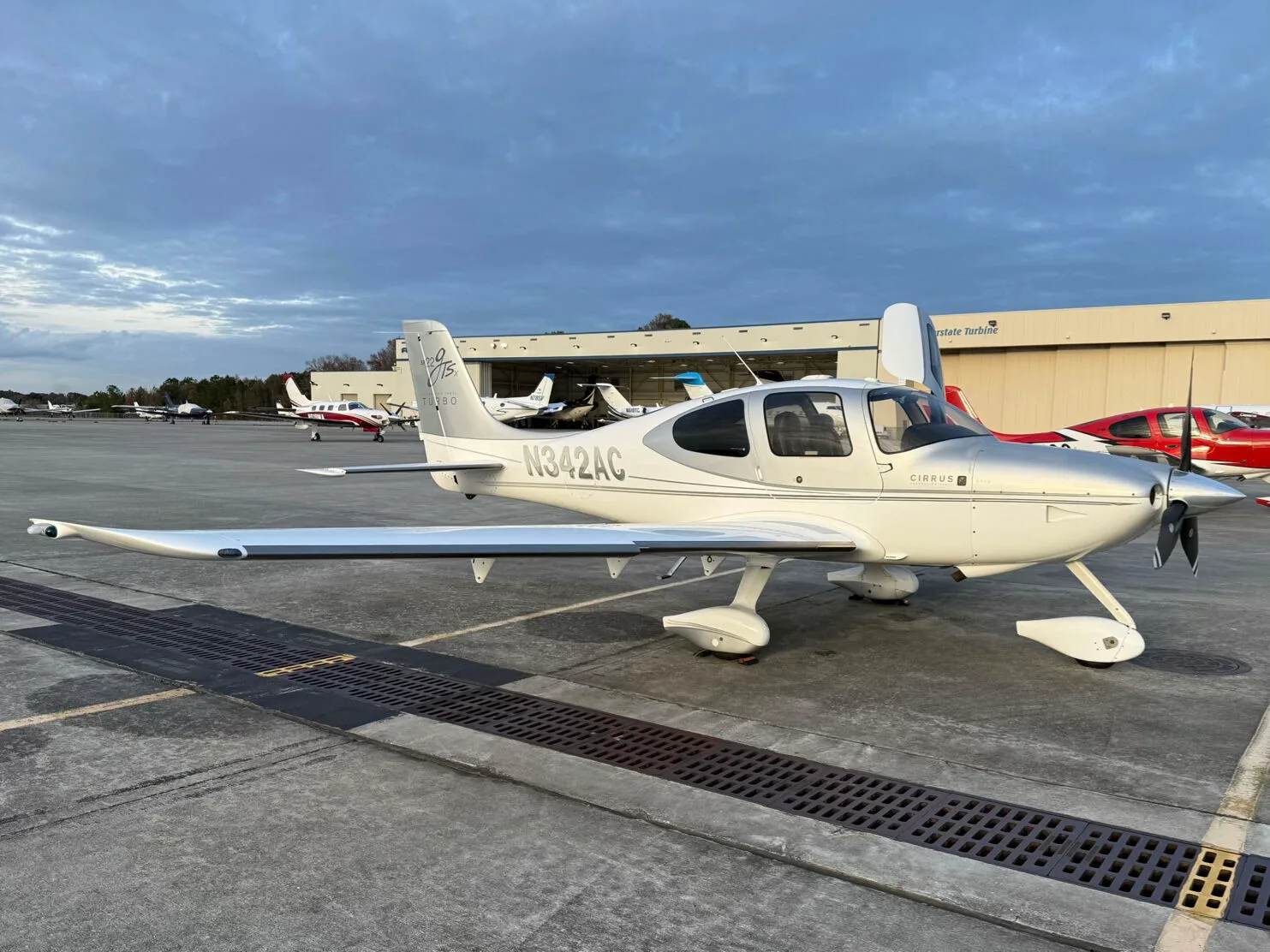 A white and silver Cirrus SR22 GTSX Turbo aircraft (N342AC) parked on an airport tarmac with hangars in the background.