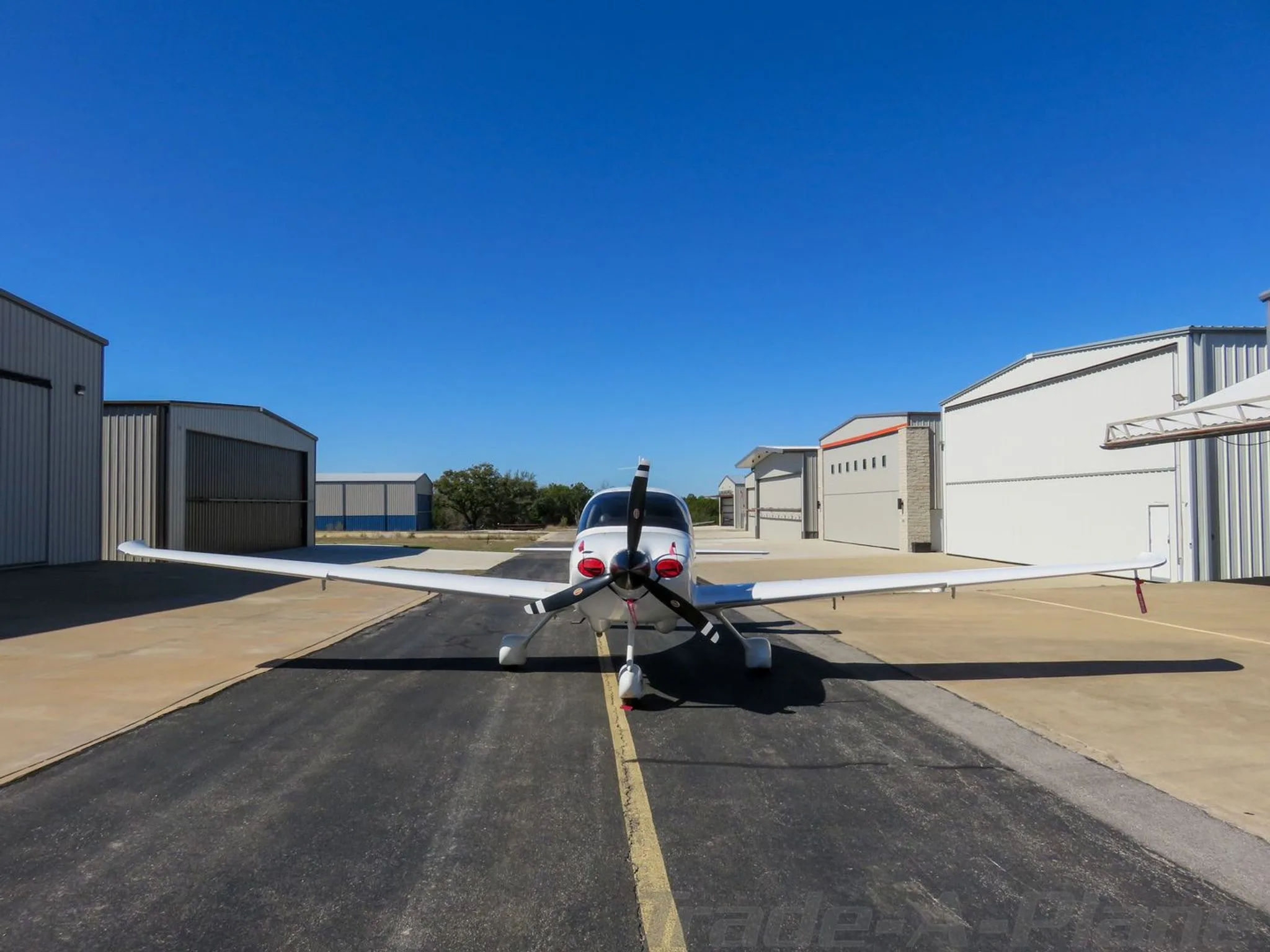 Front view of a white single-engine aircraft with a red spinner, parked on a tarmac between hangars.