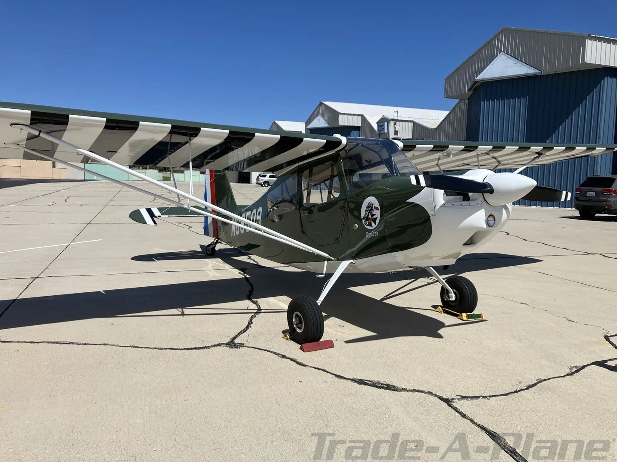A green and white single-engine aircraft with striped wings, parked on an asphalt tarmac in front of hangars.