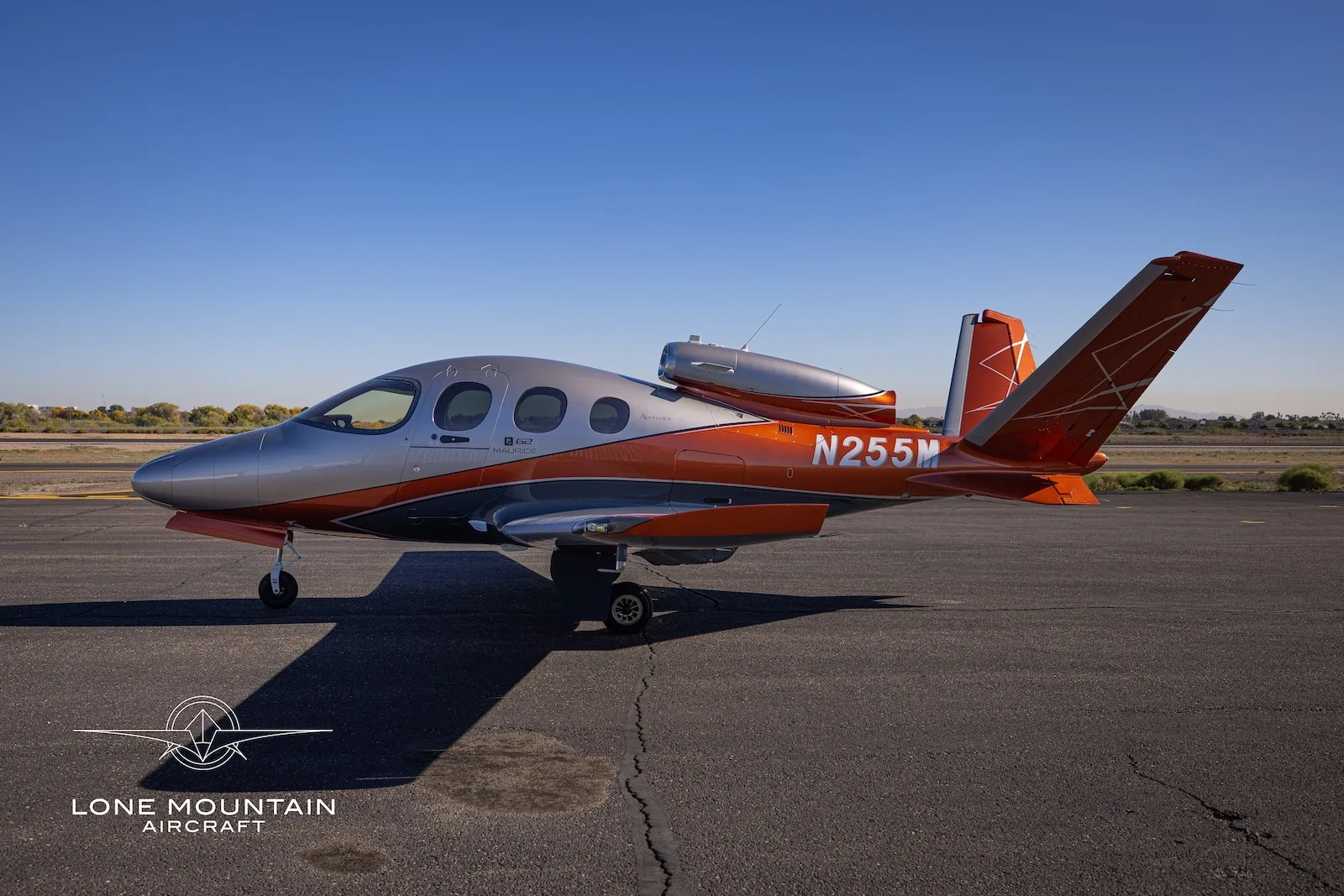 A side view of a Cirrus Vision Jet (SF50) with registration N255M, showcasing its silver and orange paint scheme on an asphalt tarmac.