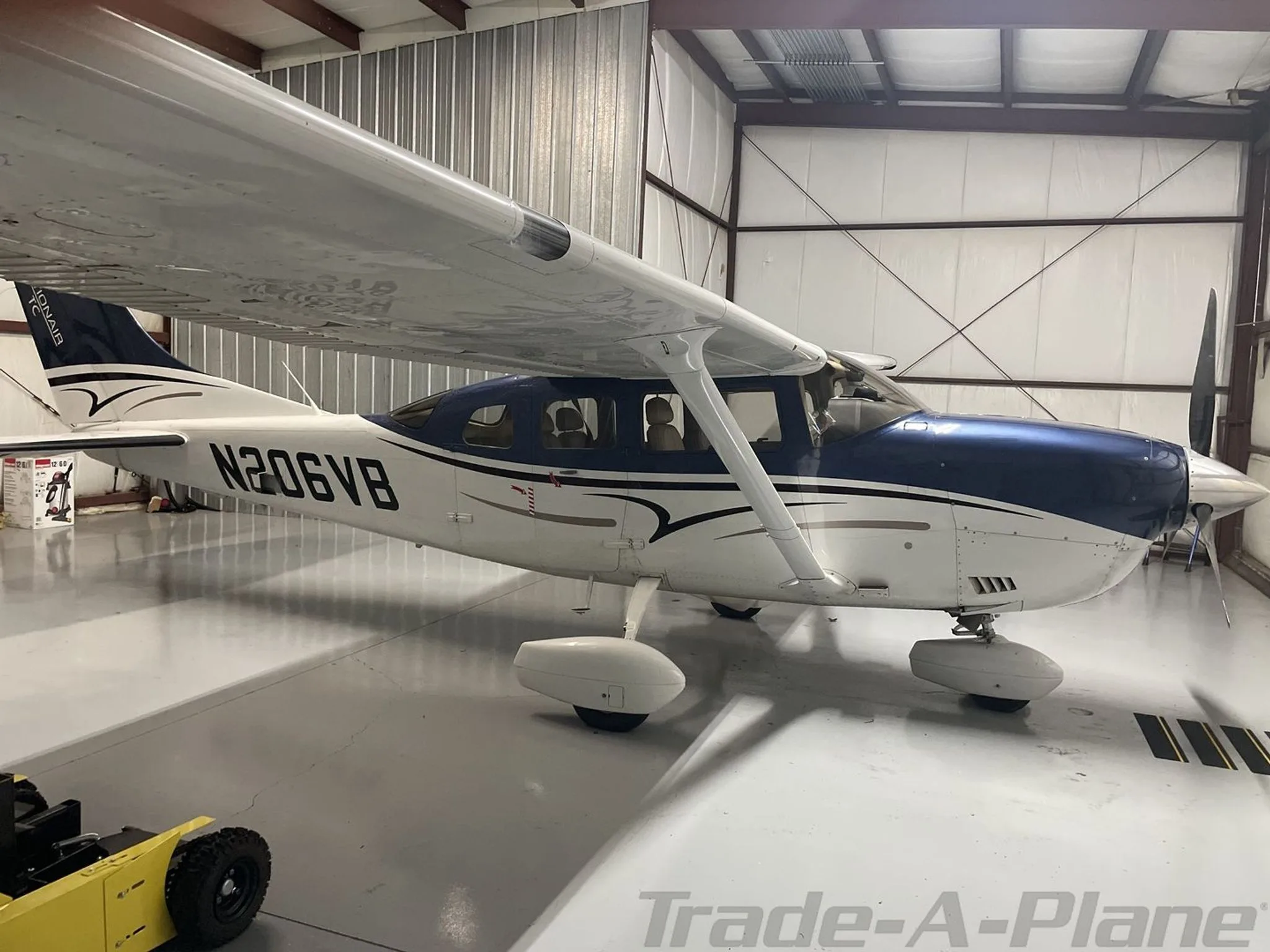 A three-quarter side view of a blue and white Cessna 182 aircraft with registration N206VB, parked in a hangar.
