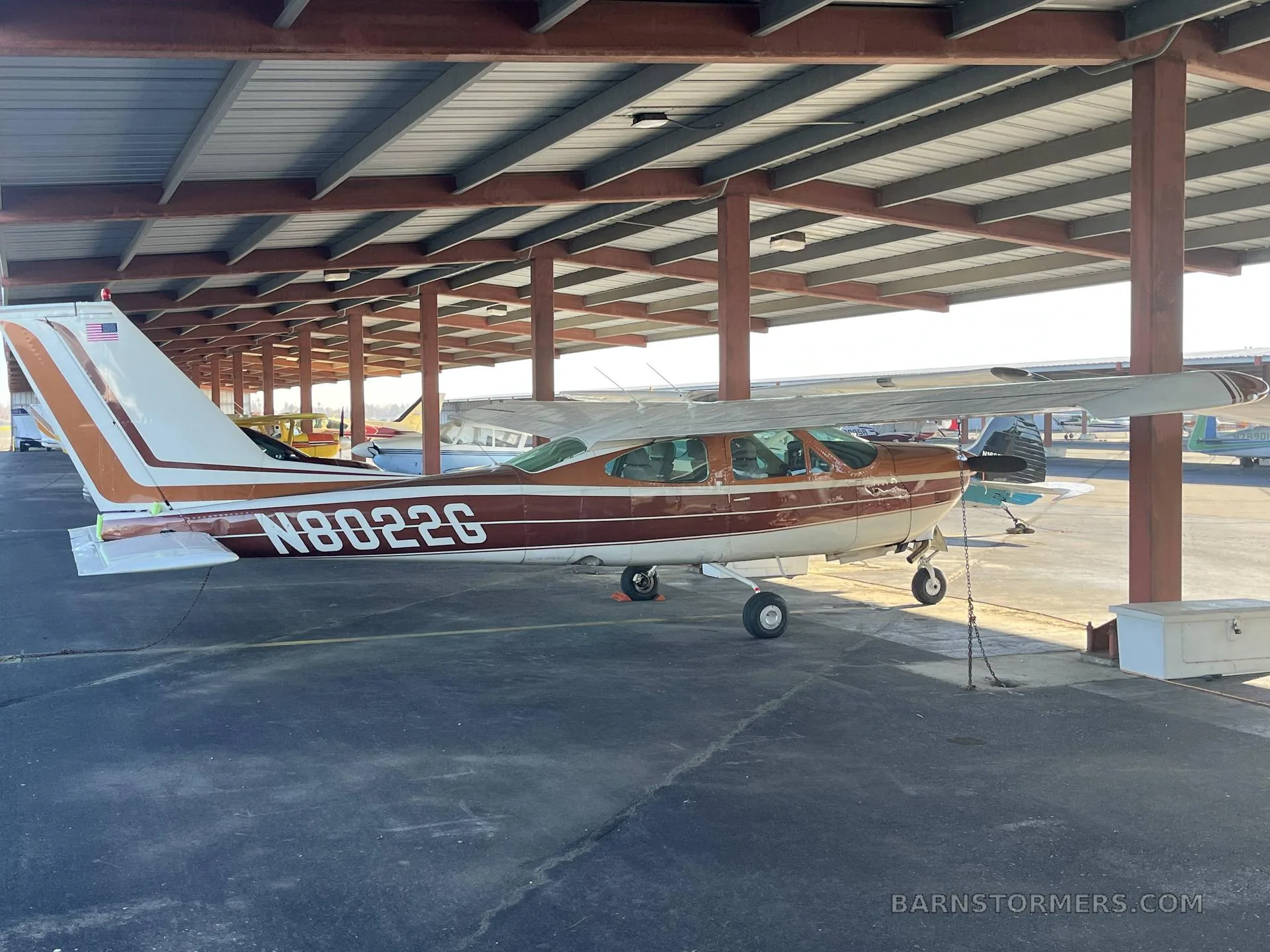 A side view of a brown and white Cessna 182 aircraft with tail number N8022G parked under a hangar roof.