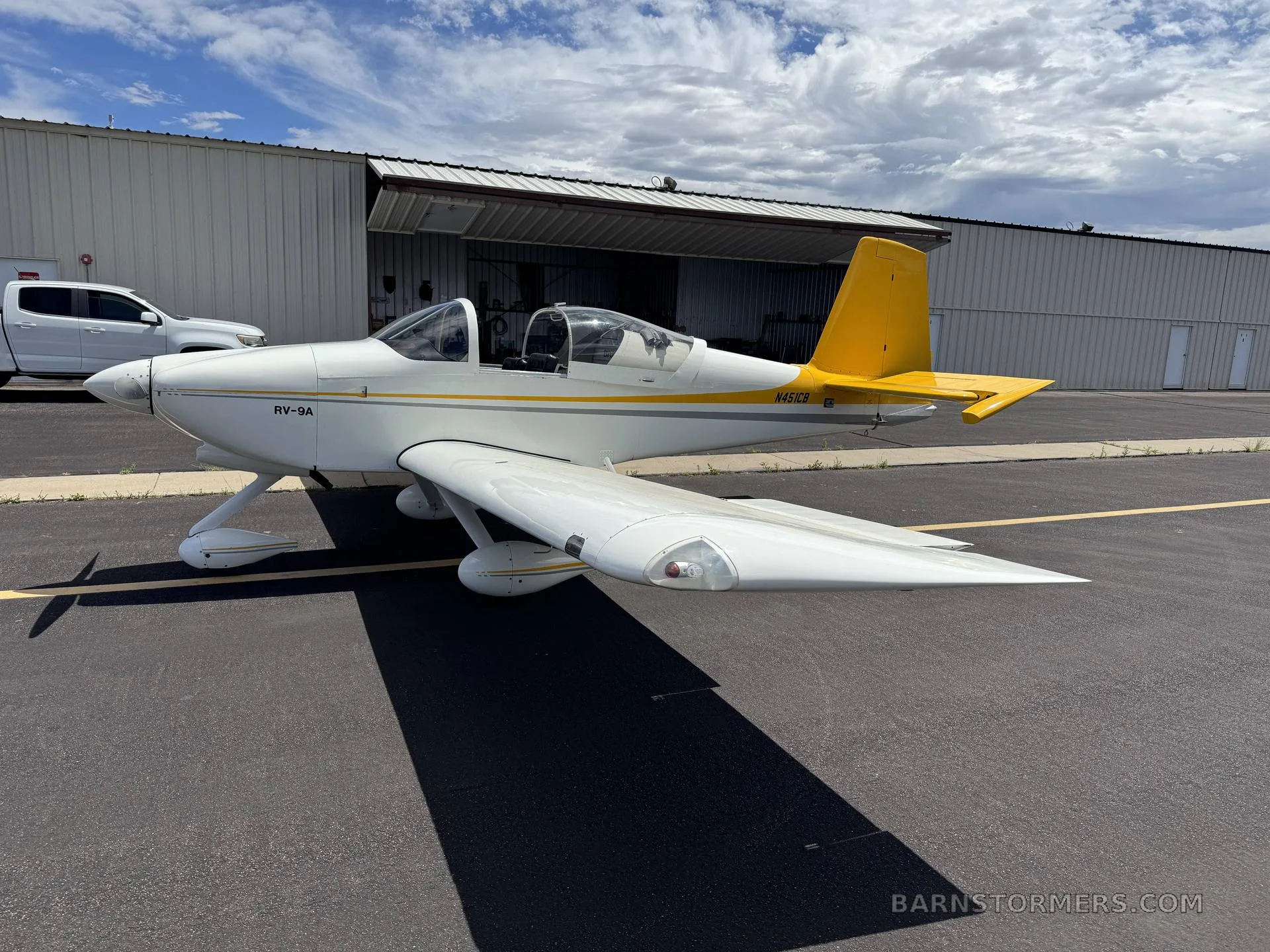 A side view of a white RV-9A aircraft with a yellow tail and stripe, parked on an asphalt ramp with a white truck in the background.