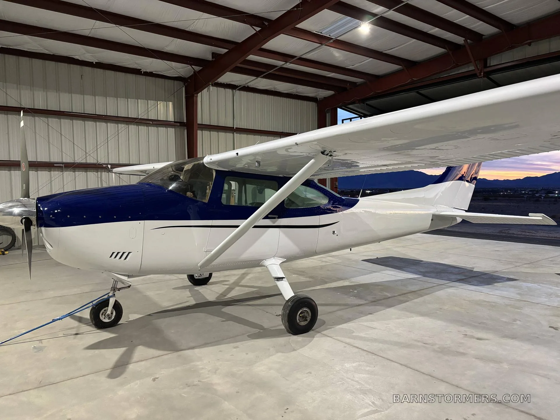 A side view of a blue and white single-engine propeller aircraft parked inside a hangar with a sunset visible outside.