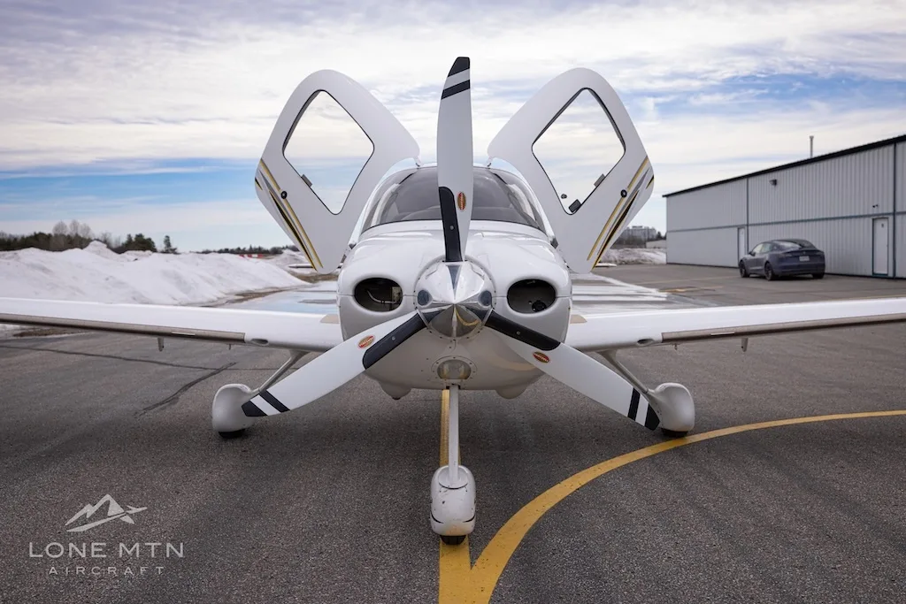A head-on view of a white Cirrus SR22 aircraft with registration C-GKKT, doors open, on an airfield.