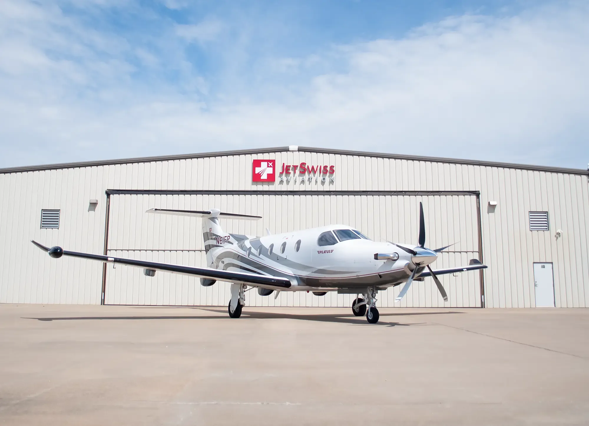 A white Pilatus PC-12 aircraft with registration N61FP is parked on the tarmac in front of a JetSwiss Aviation hangar.
