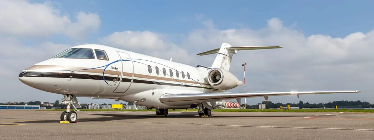 A white and brown Dassault Falcon business jet parked on an airport tarmac under a partly cloudy sky.