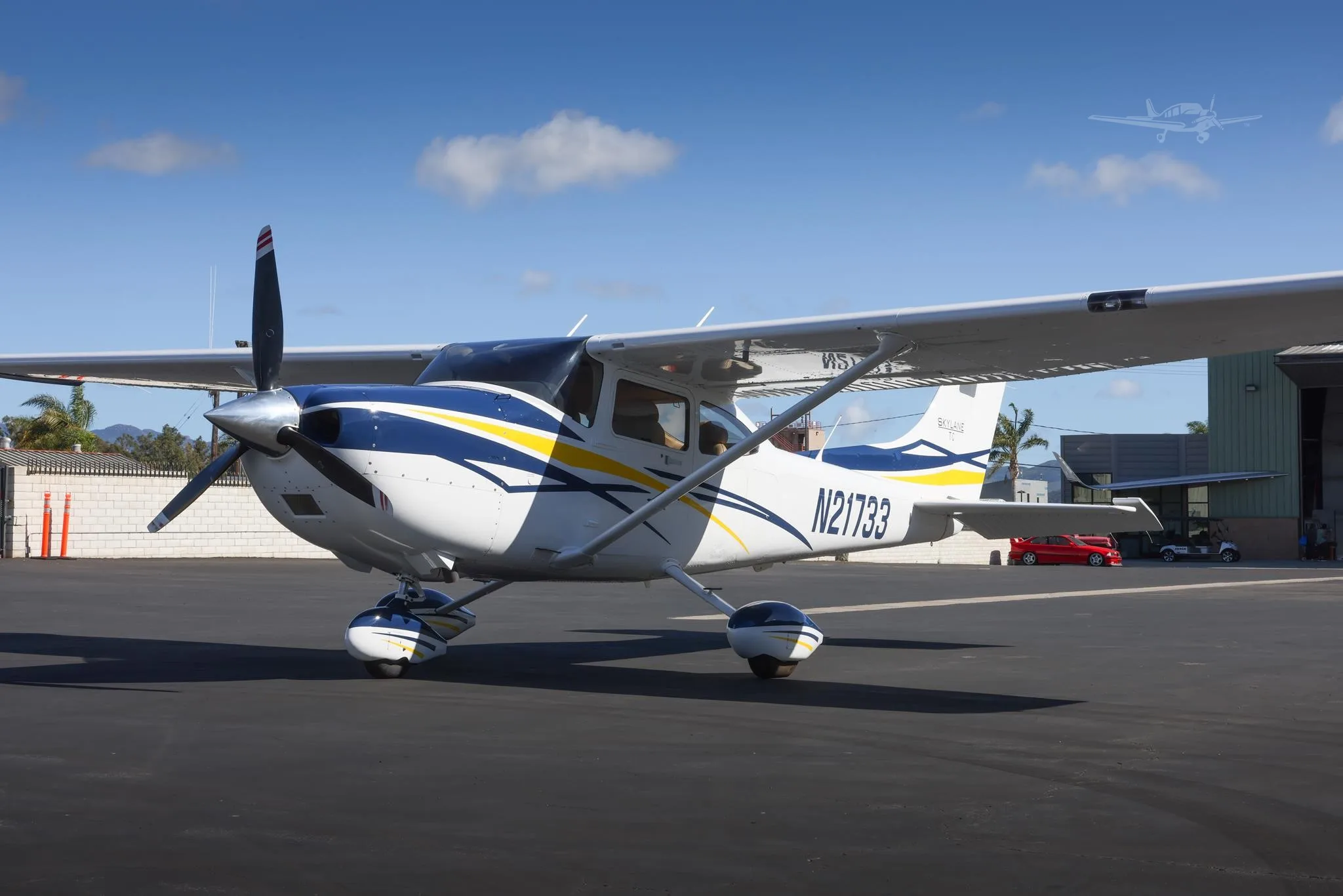 A white Cessna 182 Skylane with blue and yellow stripes is parked on an asphalt ramp at an airport under a clear blue sky.