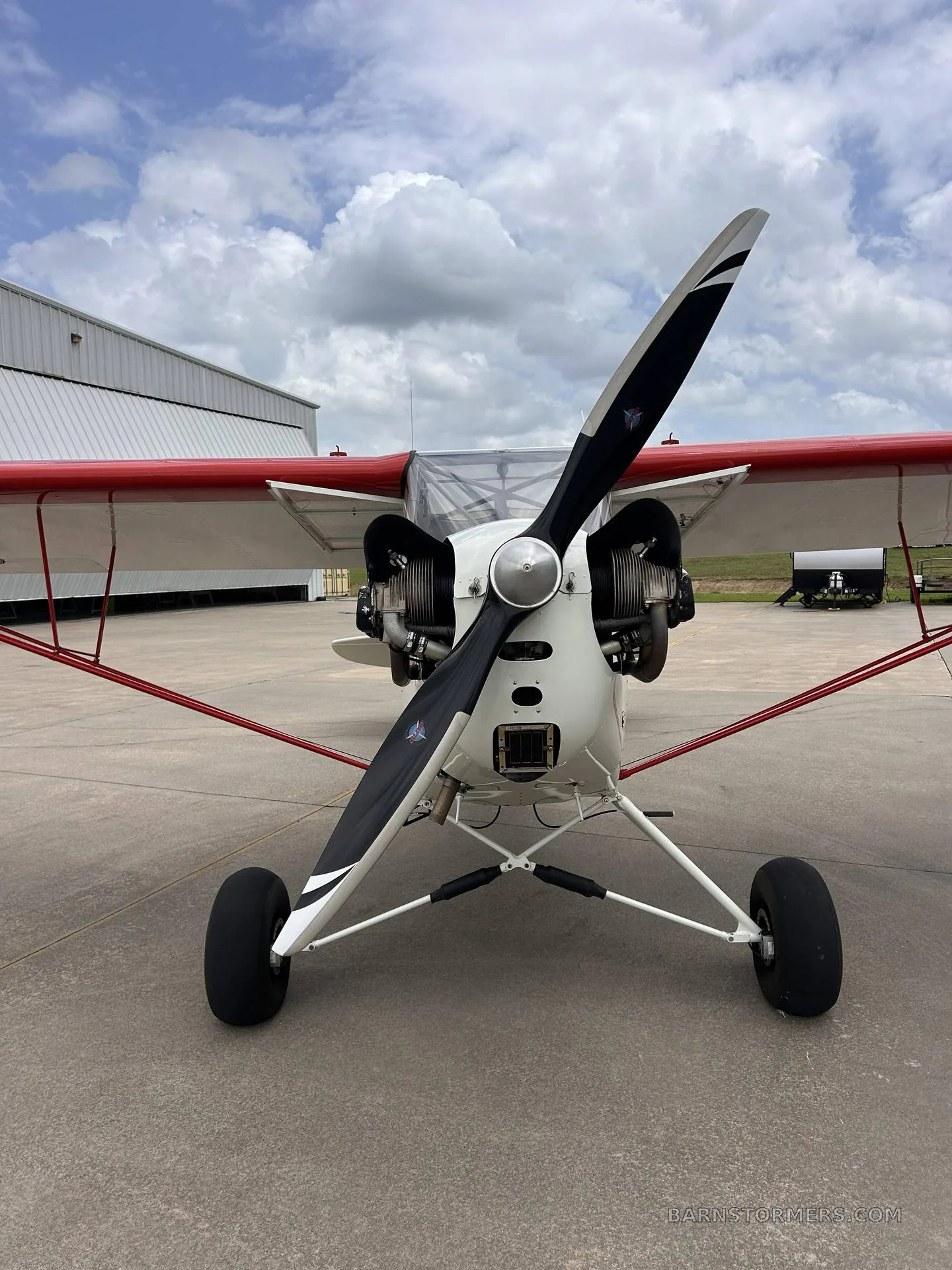 A head-on view of the white and red single-engine aircraft, showing its propeller, exposed engine, and landing gear.