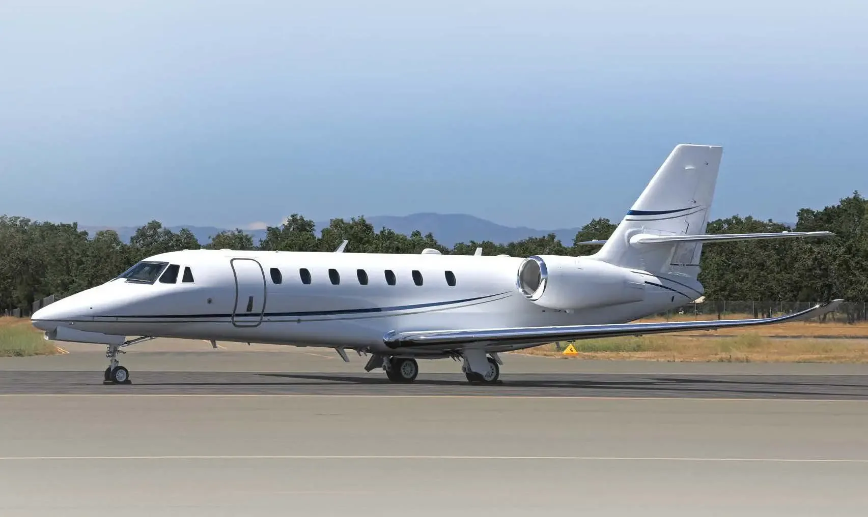 A white business jet with blue accent stripes parked on an airfield with trees and mountains in the background.