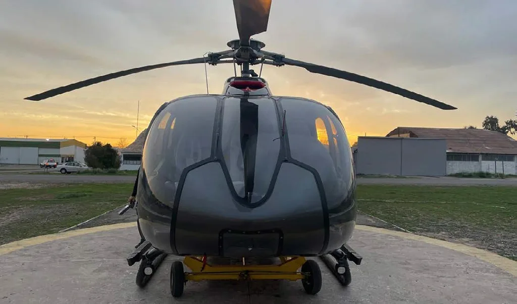 A dark grey and red helicopter is viewed from the front on a helipad at sunset.