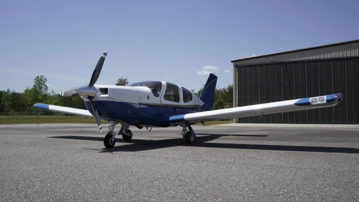 A front-left view of a white and blue single-engine aircraft with a propeller on an airfield.