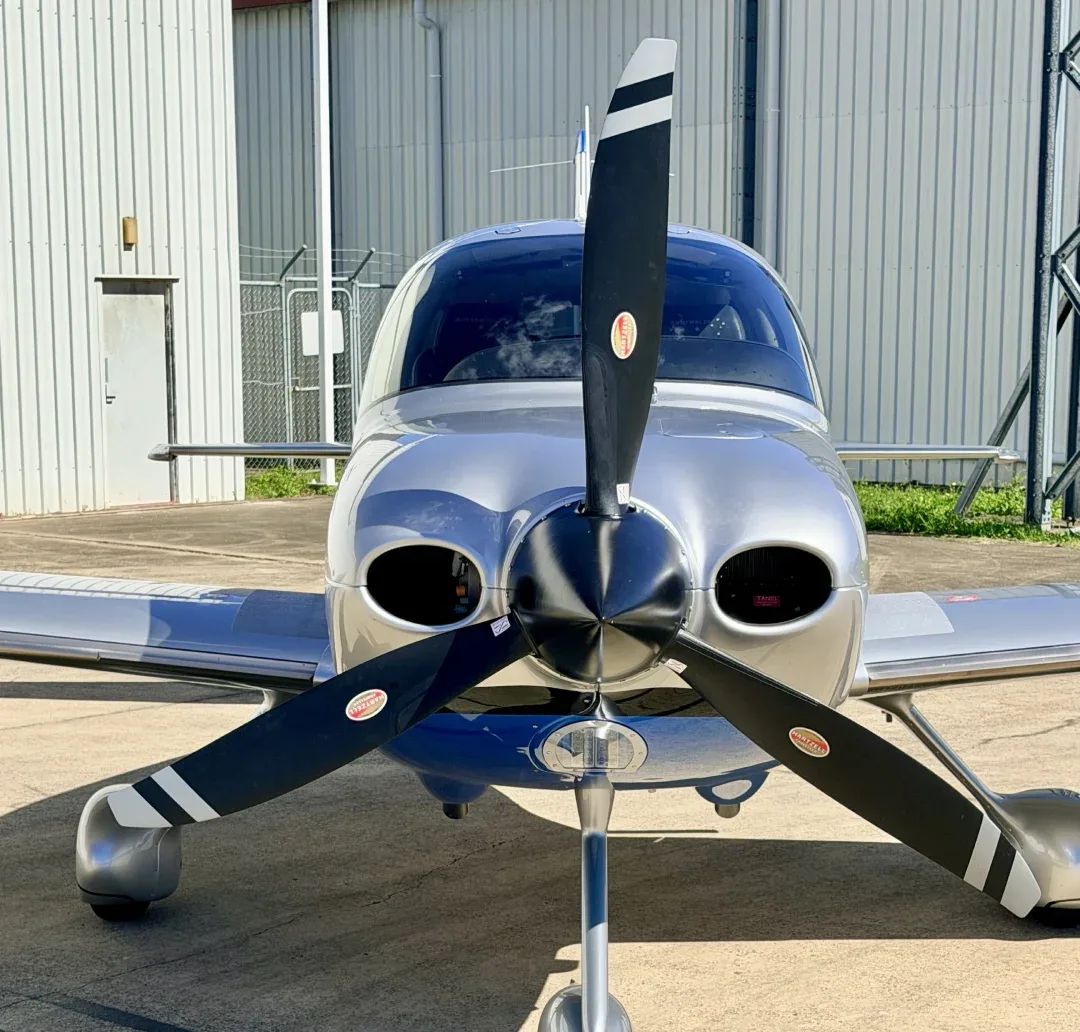 A silver single-engine propeller aircraft with black and white striped blades, viewed from the front on an airport tarmac.