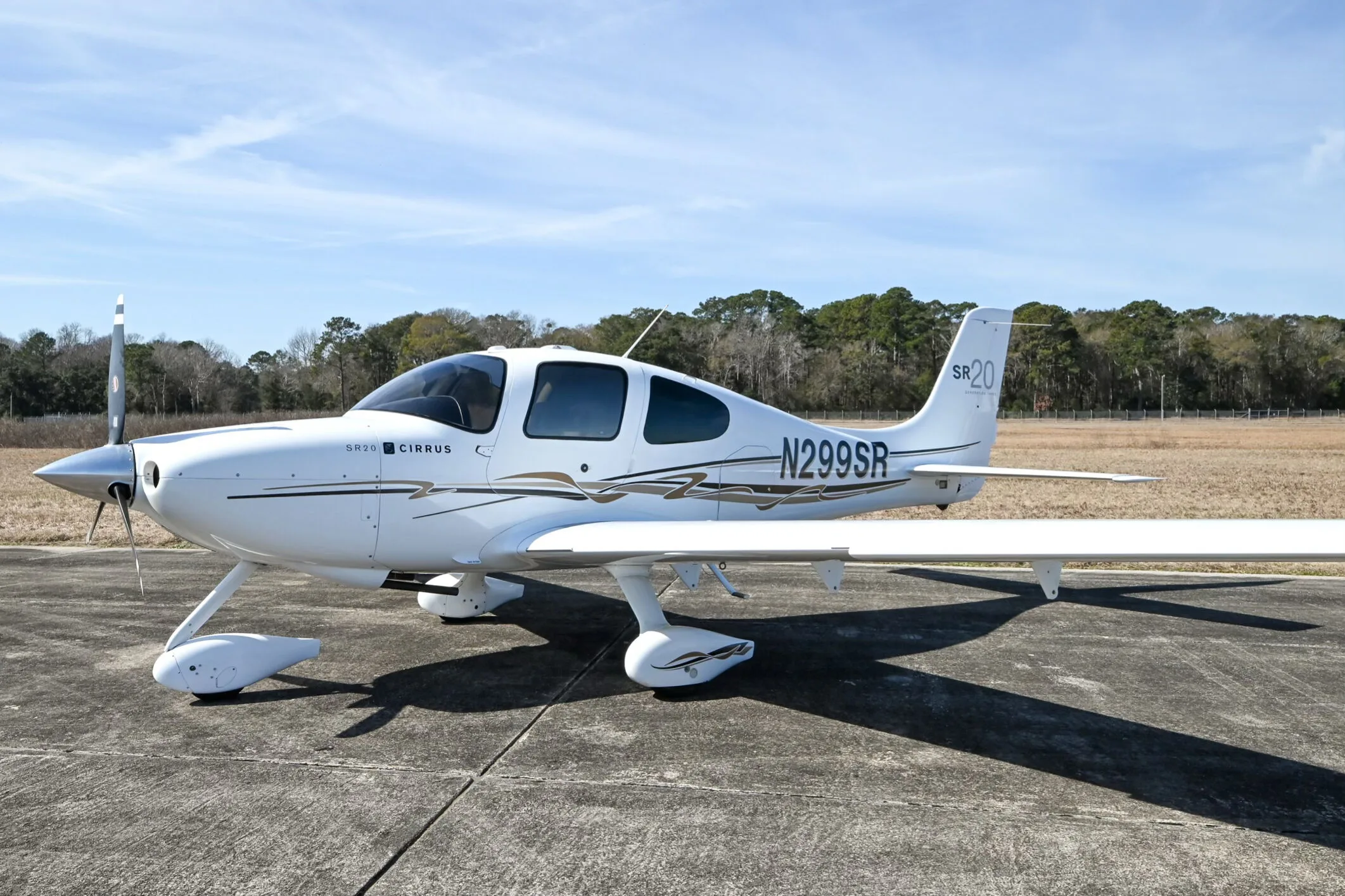 A white Cirrus SR20 aircraft with black and gold accent stripes, registration N299SR, parked on an airfield with trees in the background.