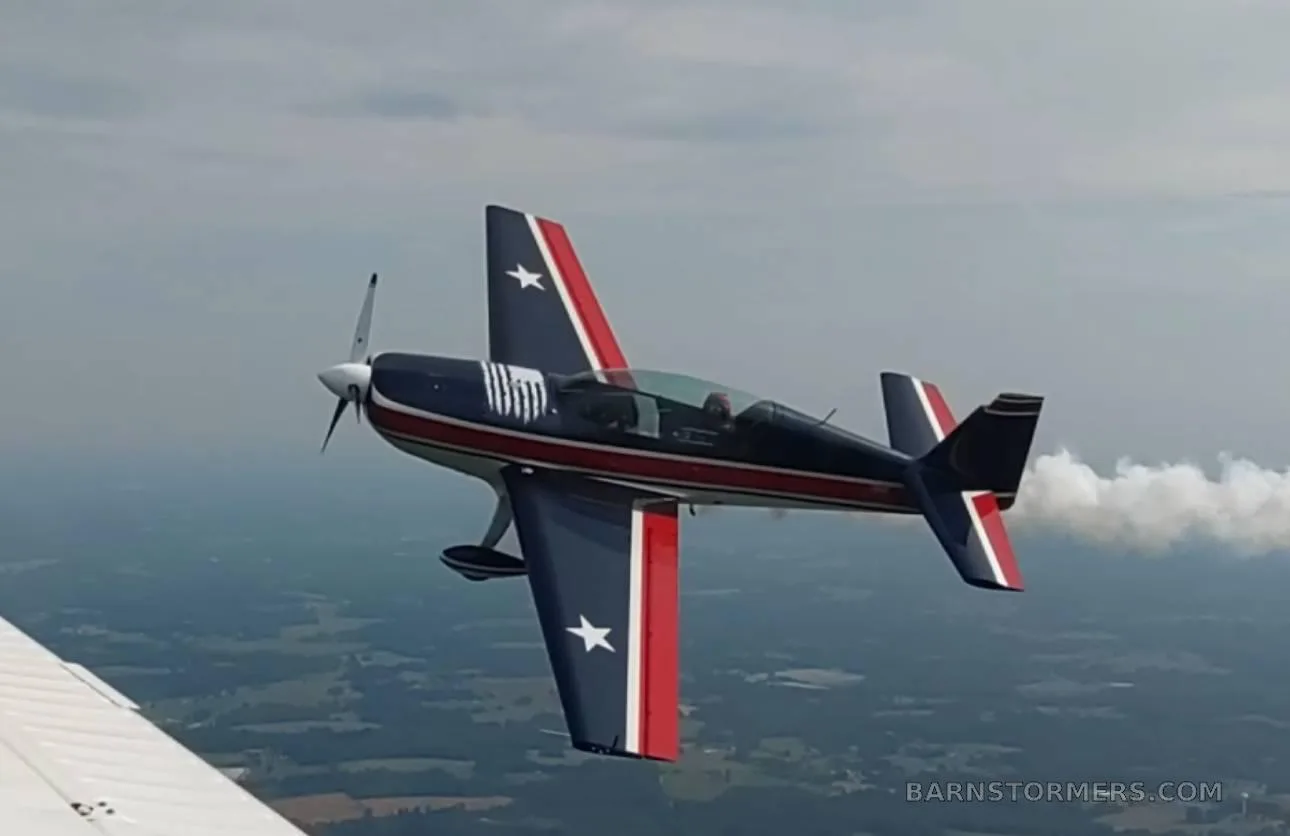 A dark blue, red, and white aerobatic aircraft with stars on its wings and tail, trailing smoke during a maneuver over a green landscape.