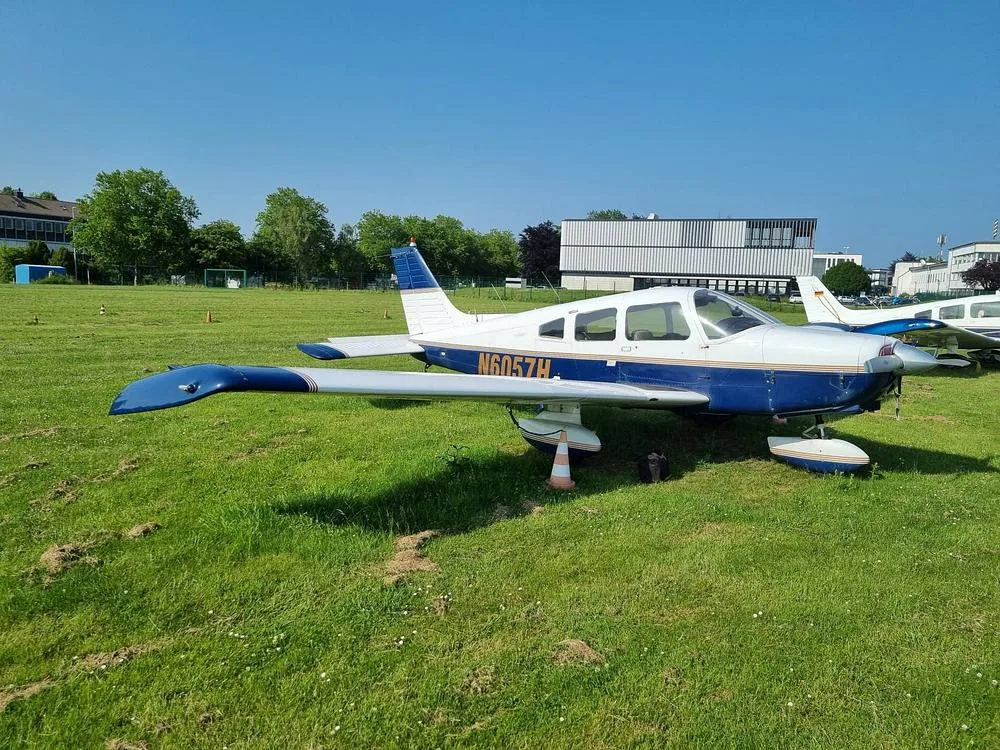 A white and blue Piper PA-28 Cherokee aircraft, registration N6057H, parked on a grassy airfield under a clear blue sky.