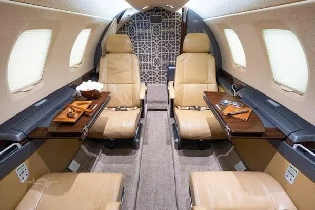 The luxurious tan leather interior of a private jet cabin with fold-out tables and patterned bulkhead.