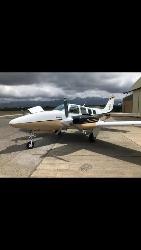 A white and gold twin-engine propeller aircraft, possibly a Beechcraft Baron, parked on an asphalt ramp under a cloudy sky.