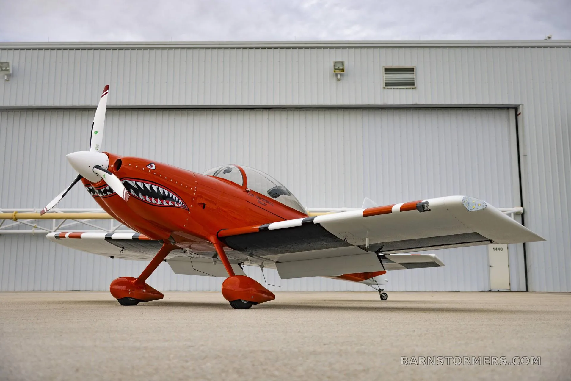 An orange and white single-engine aircraft with a shark mouth paint scheme on the nose, parked in front of a hangar.