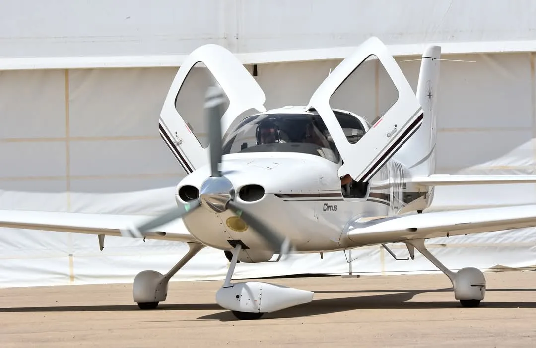 A white Cirrus single-engine aircraft with dark stripes, propeller spinning and doors open, sits on a tarmac with a white structure in the background.