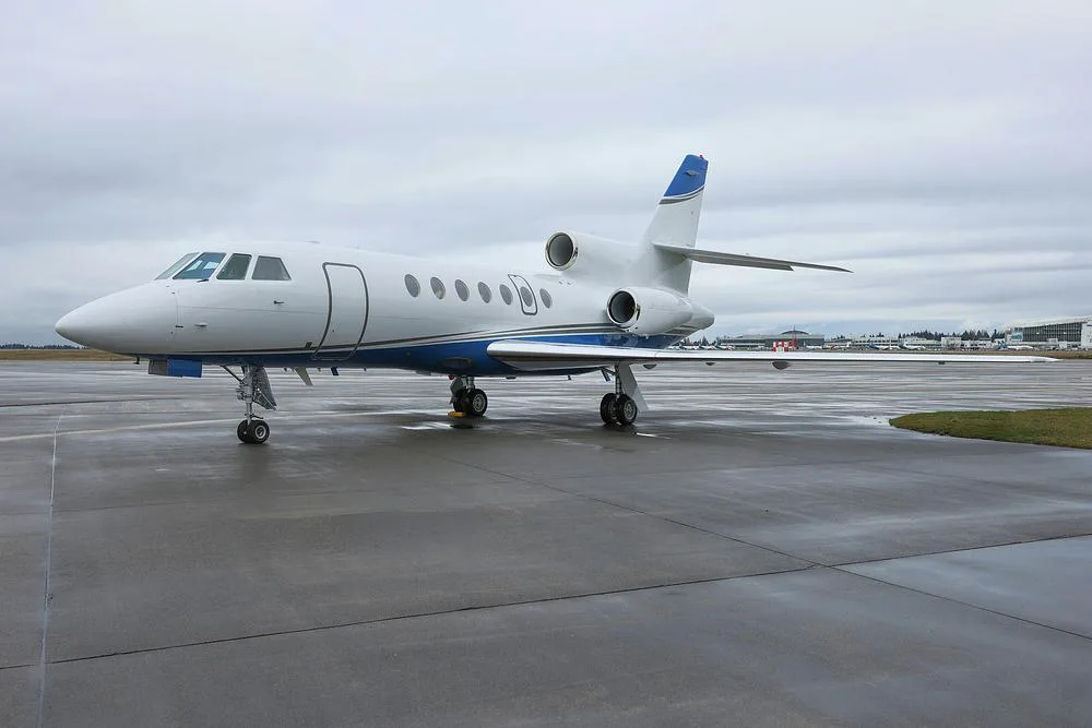 A white and blue Dassault Falcon 900EX business jet parked on a wet tarmac under a cloudy sky.