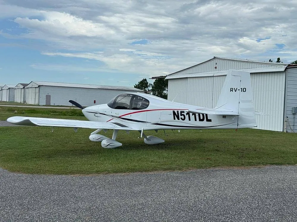 A white Van's RV-10 aircraft with red and black accents, registration N511DL, parked on grass next to a line of hangars.