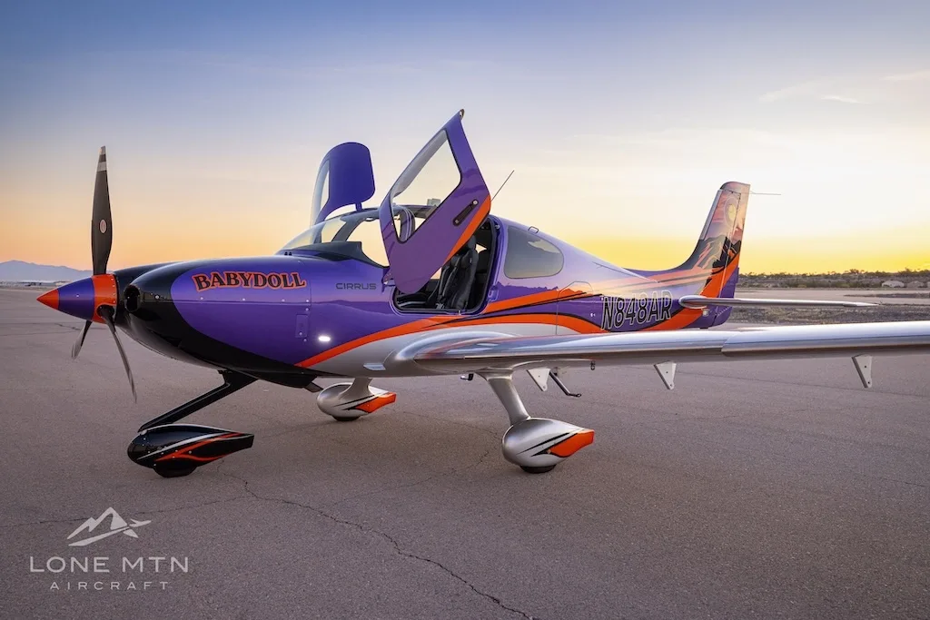 Three-quarter front view of a purple and silver Cirrus SR22T with open doors, featuring a Colorado flag tail design, parked on a tarmac at sunset.