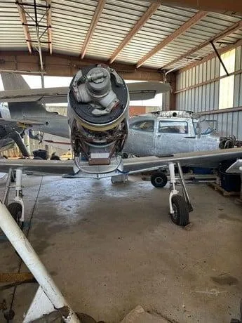 A single-engine propeller aircraft with its engine cowling open, parked in a hangar with another aircraft visible in the background.