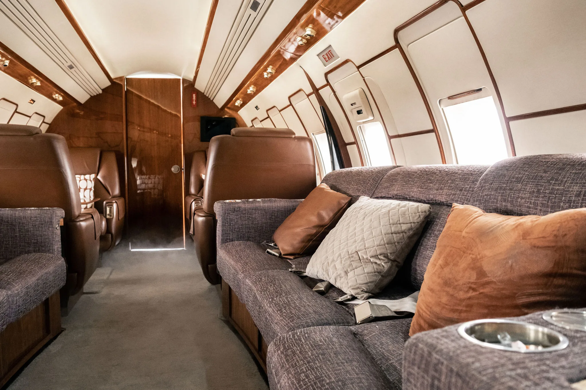 A private jet cabin interior with a grey sofa, pillows, and brown leather seats leading to the rear.
