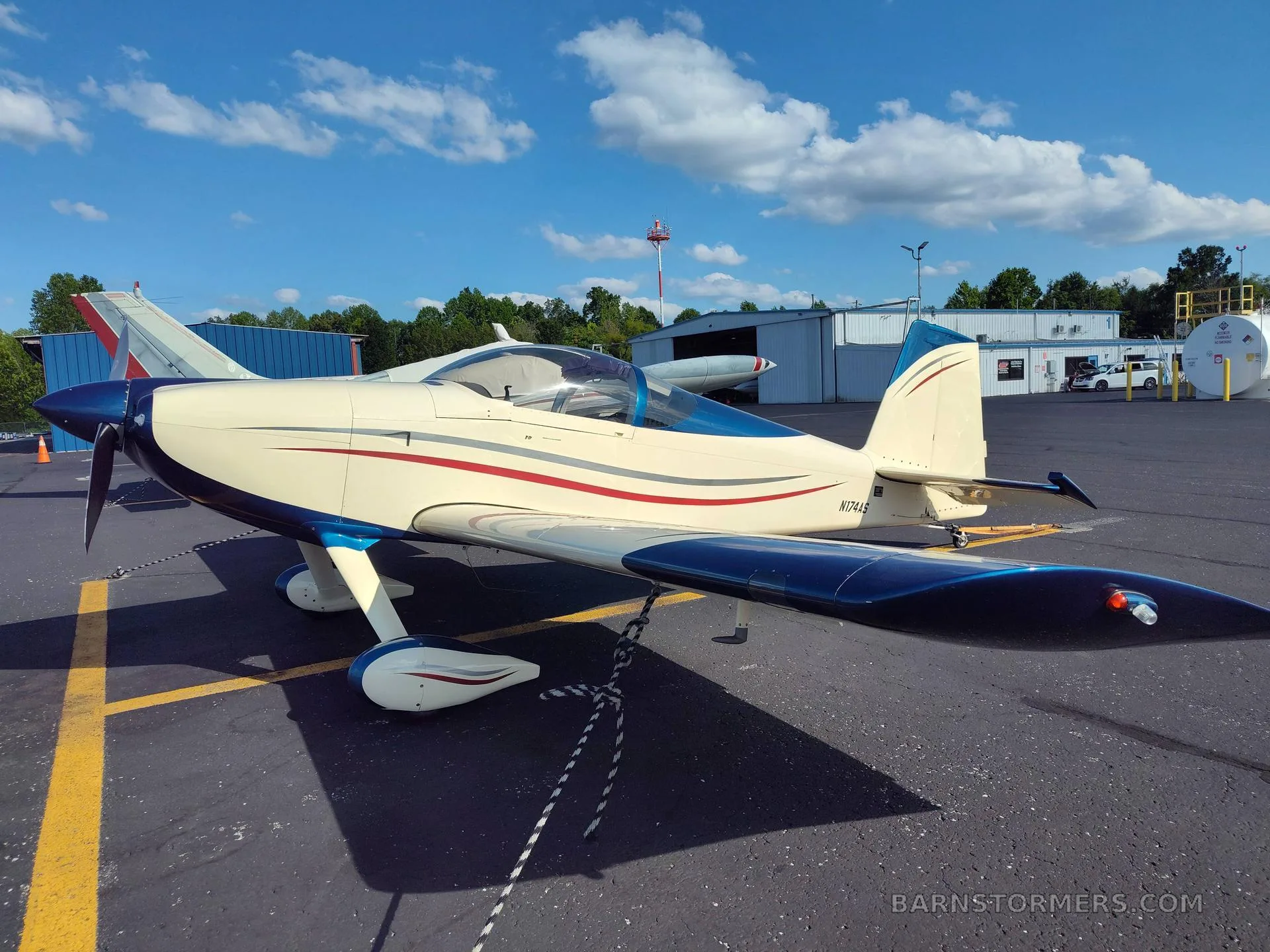 A cream and blue single-engine aircraft with red and grey stripes, parked on an asphalt tarmac under a blue sky.