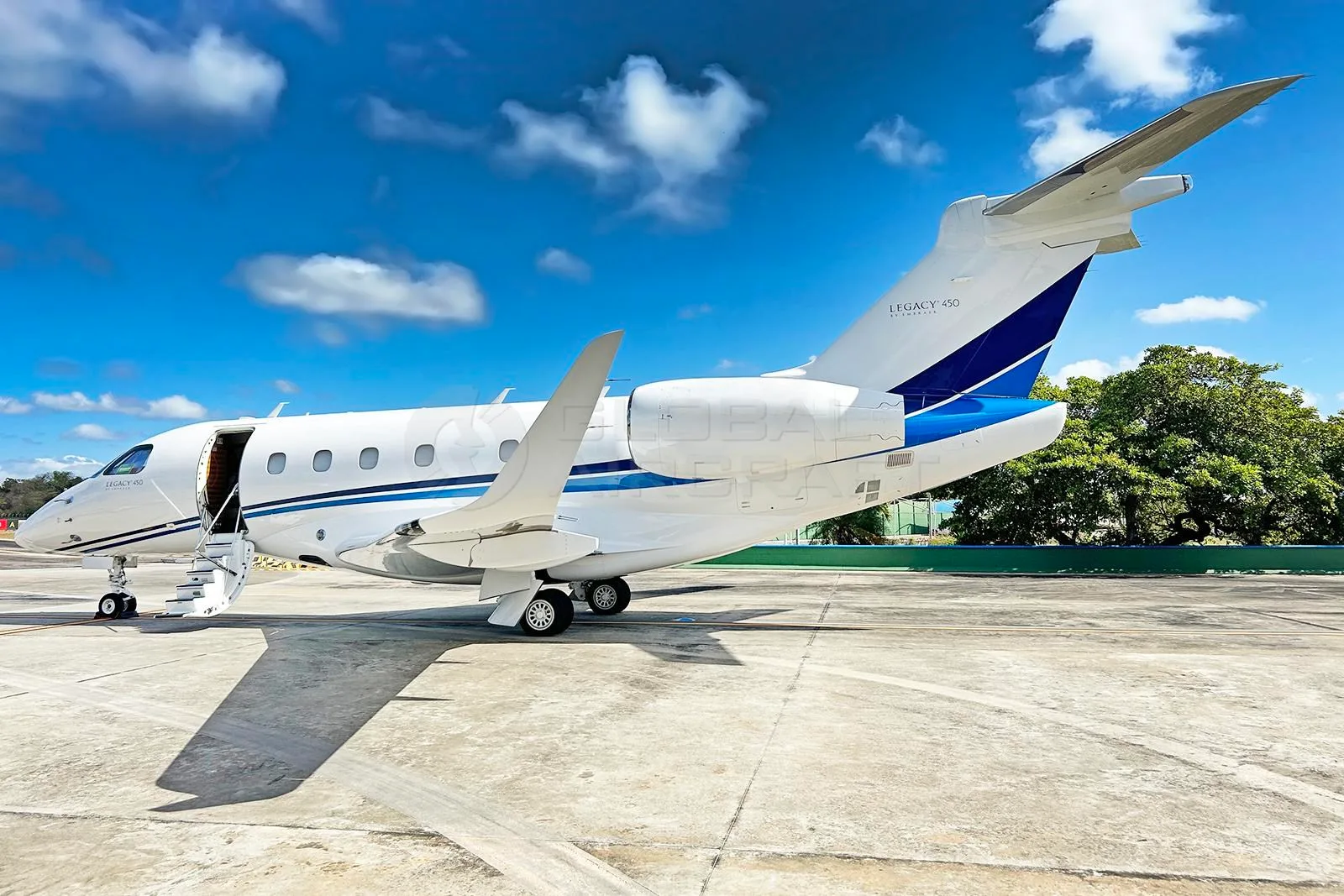 A white Embraer Legacy 450 business jet with blue tail stripes and an open passenger door on an airport tarmac.