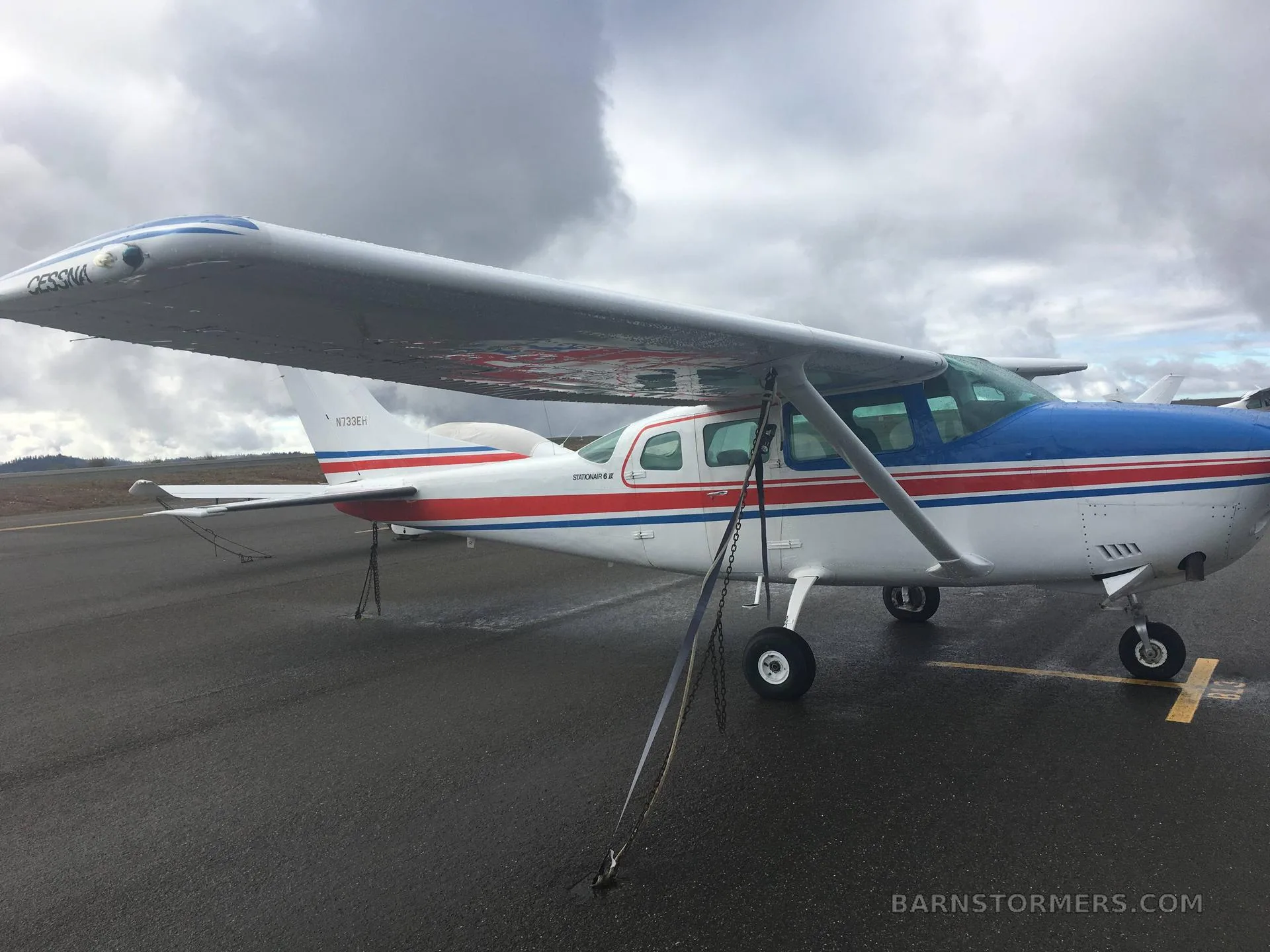 A white, red, and blue Cessna Stationair 6 II (N733EH) parked on a wet tarmac under a cloudy sky.