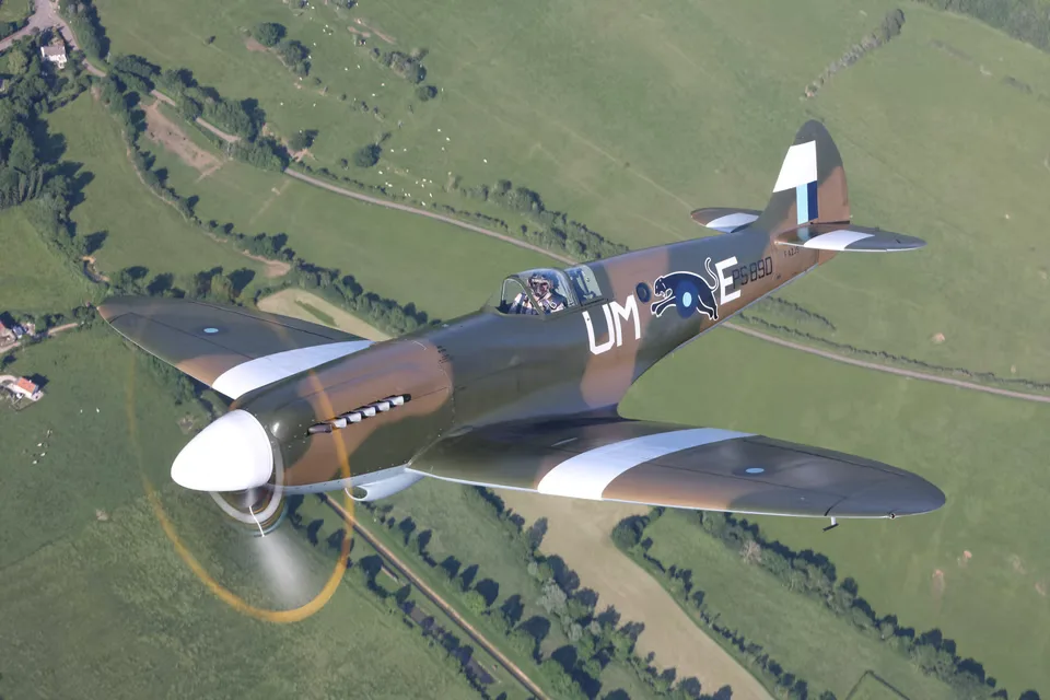 A Supermarine Spitfire in camouflage livery flying over a lush green, rural landscape.
