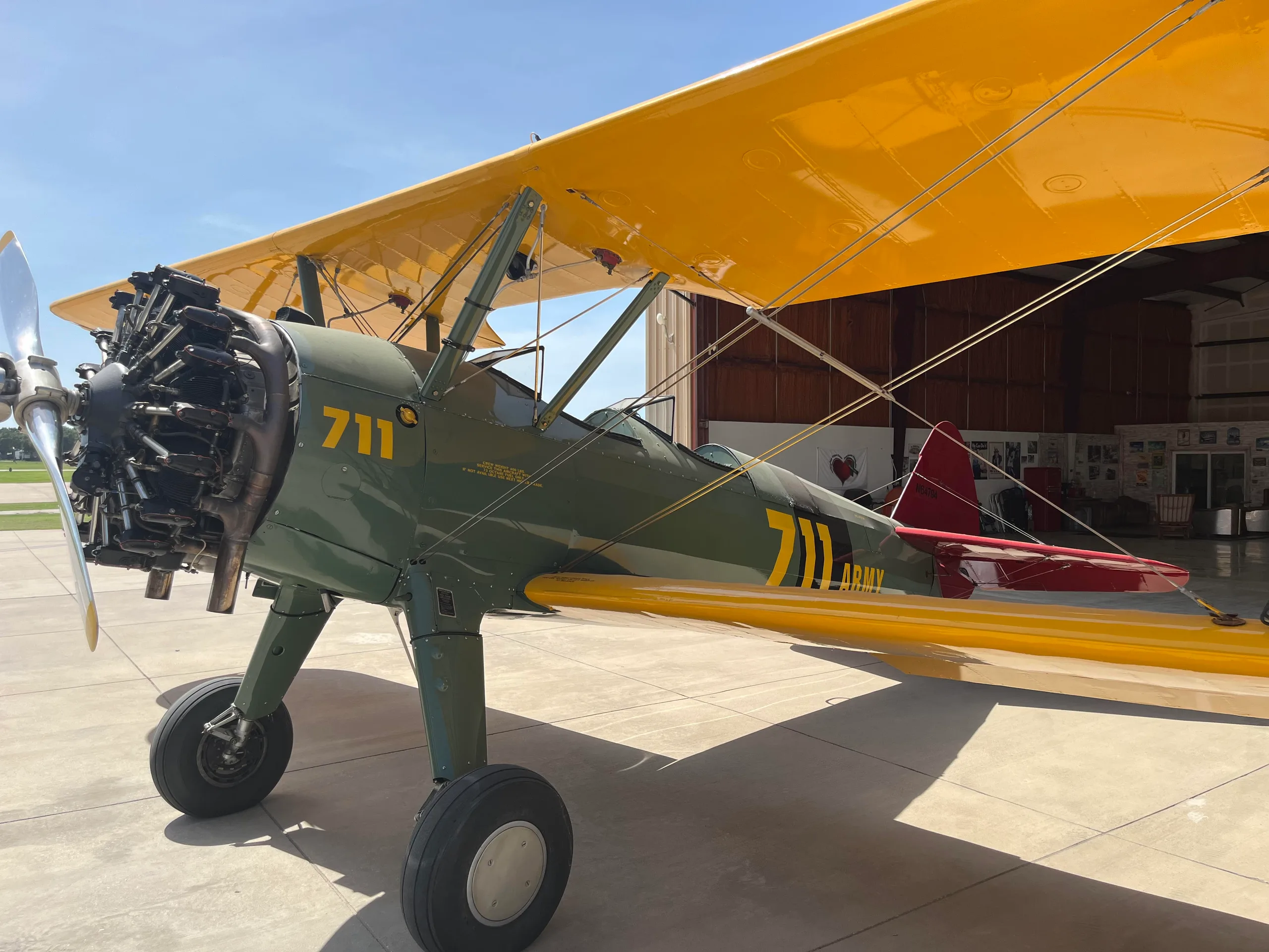 A green biplane with yellow wings and '711 ARMY' on its fuselage is parked on a tarmac with a hangar in the background.