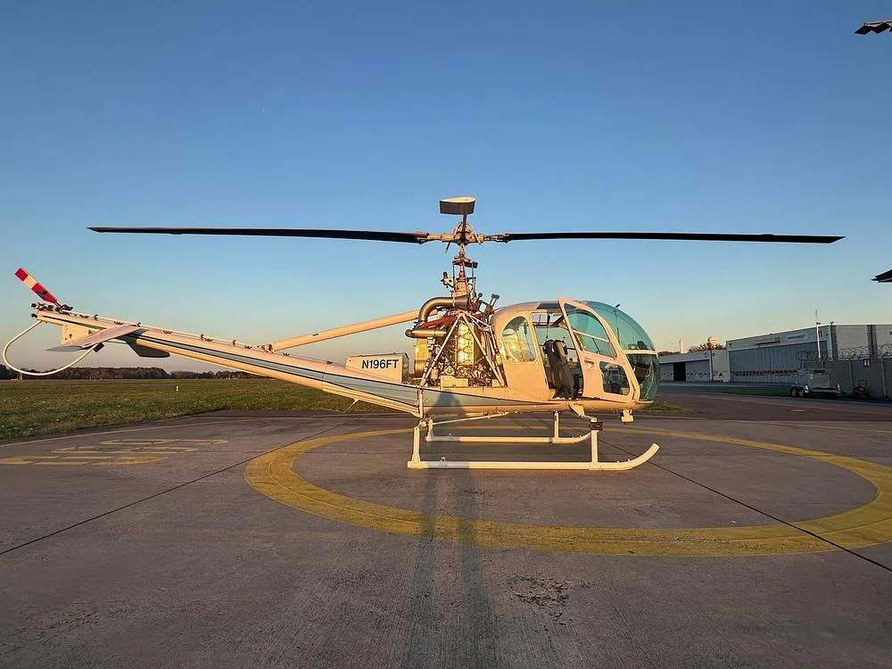 A white Hiller UH-12 helicopter with registration N196FT is parked on an airfield under a clear sky.