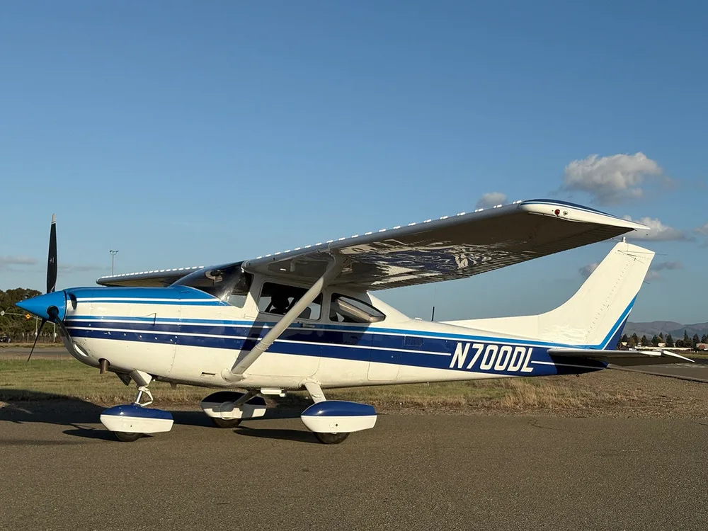 A white and blue Cessna 182 Skylane (N700DL) parked on an airfield under a clear sky.