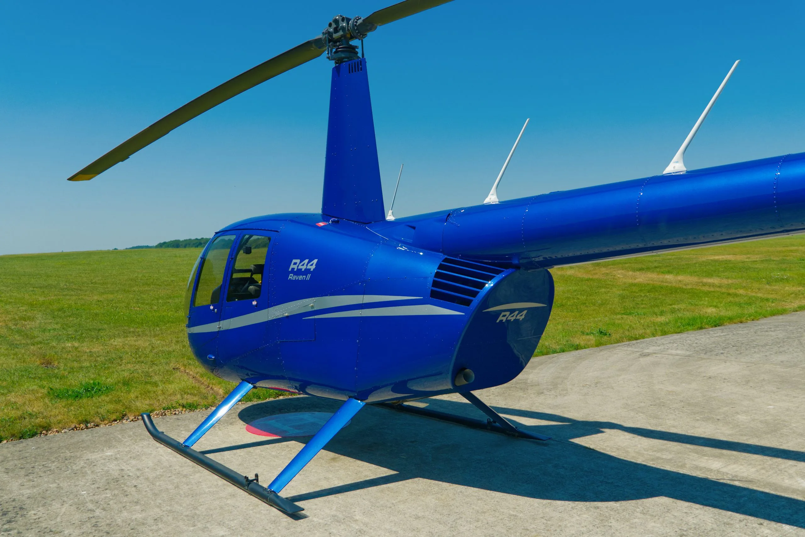 A blue Robinson R44 Raven II helicopter with a white stripe sits on a concrete pad next to a grassy field under a clear sky.