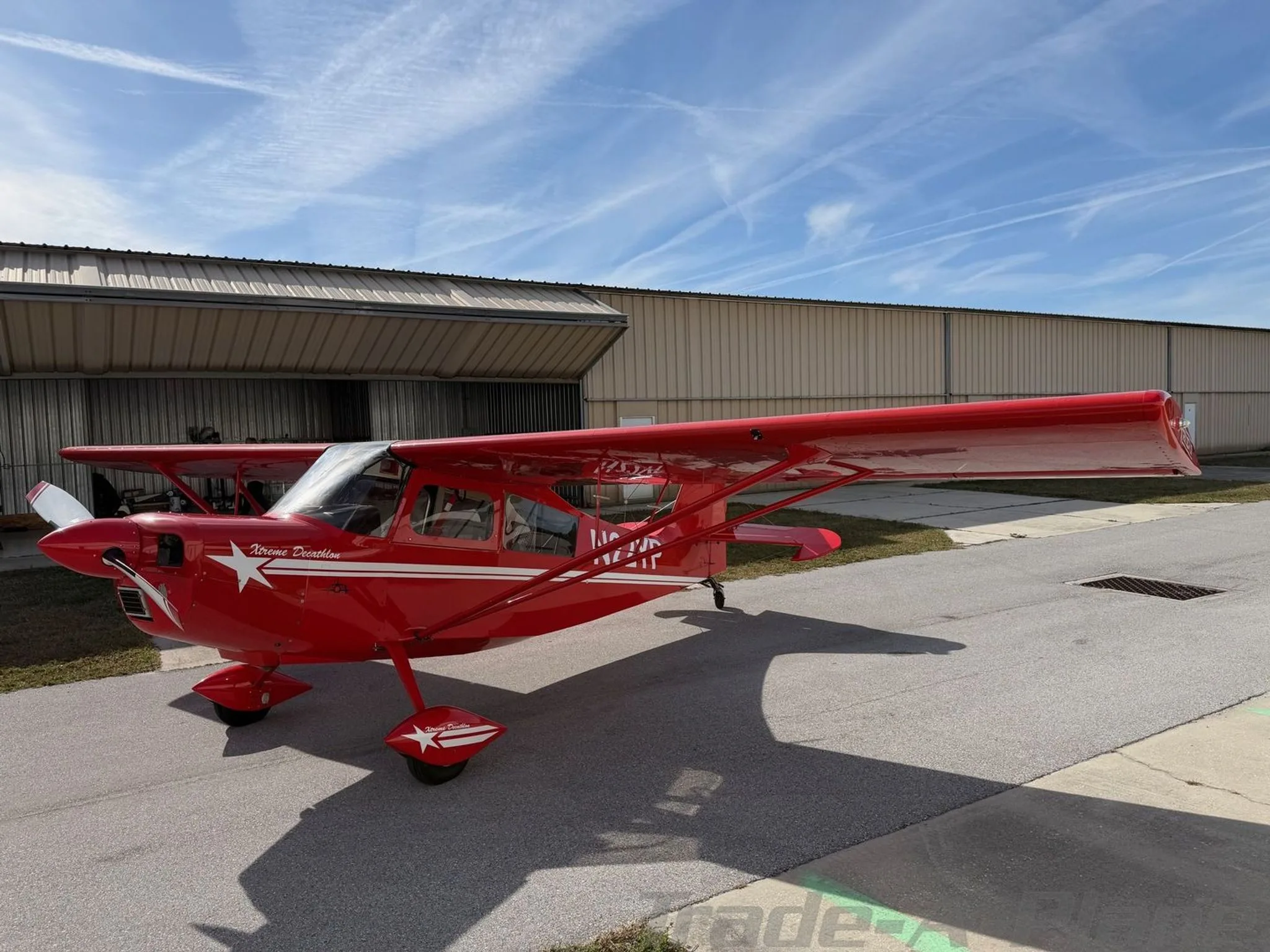 A red American Champion Xtreme Decathlon aircraft with white stripes and a star logo parked on an asphalt ramp next to a hangar.
