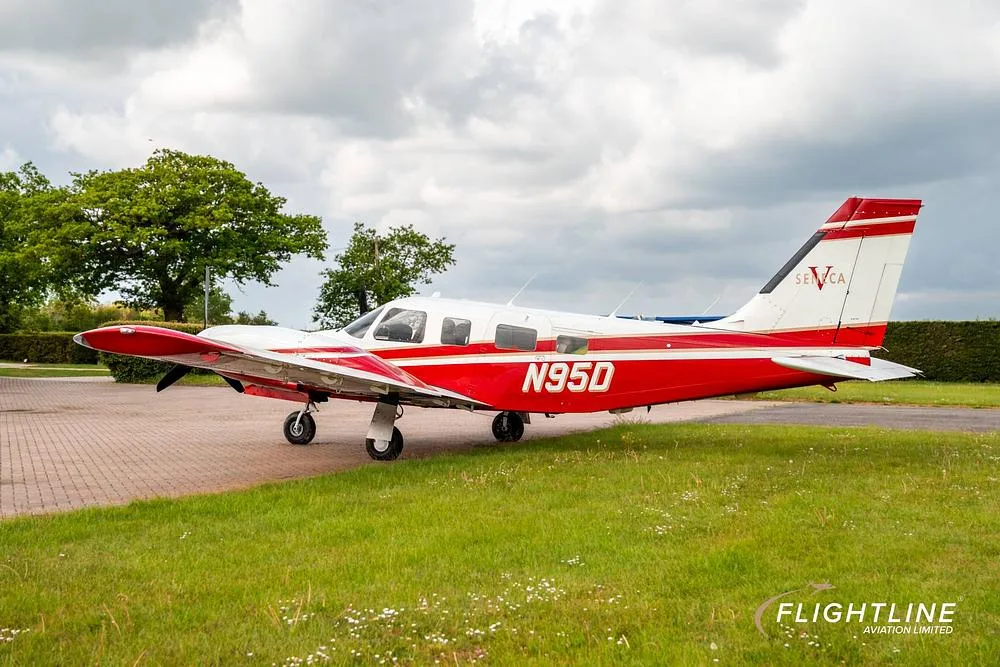 A red and white Piper Seneca aircraft with registration N95D parked on a paved surface, viewed from the left side with trees in the background.
