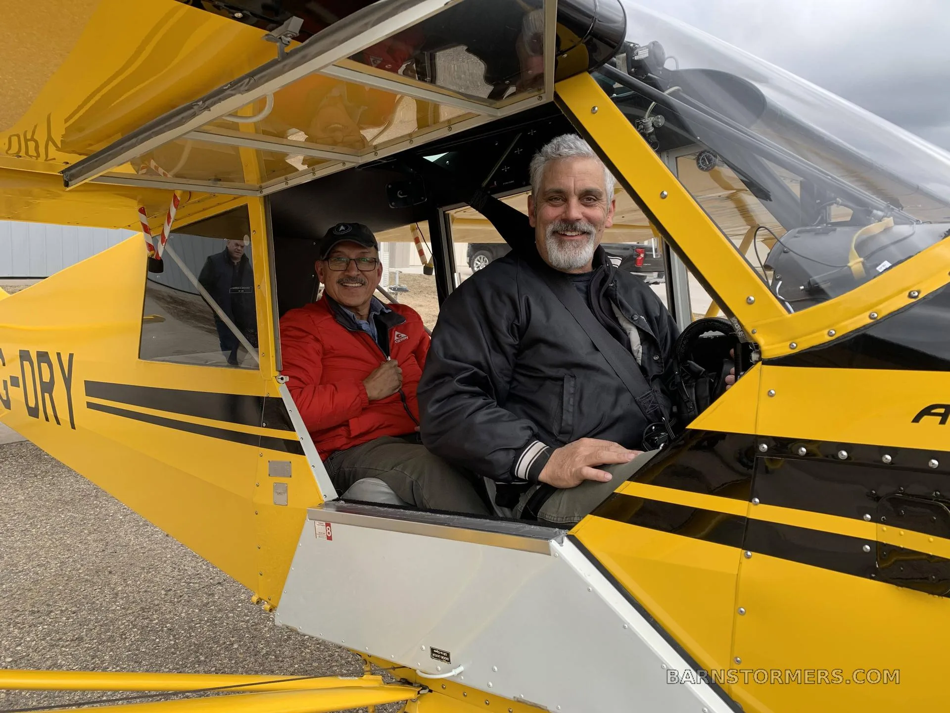 Two men smiling while seated in the open cockpit of a yellow and black Aviat Husky A-1 aircraft.