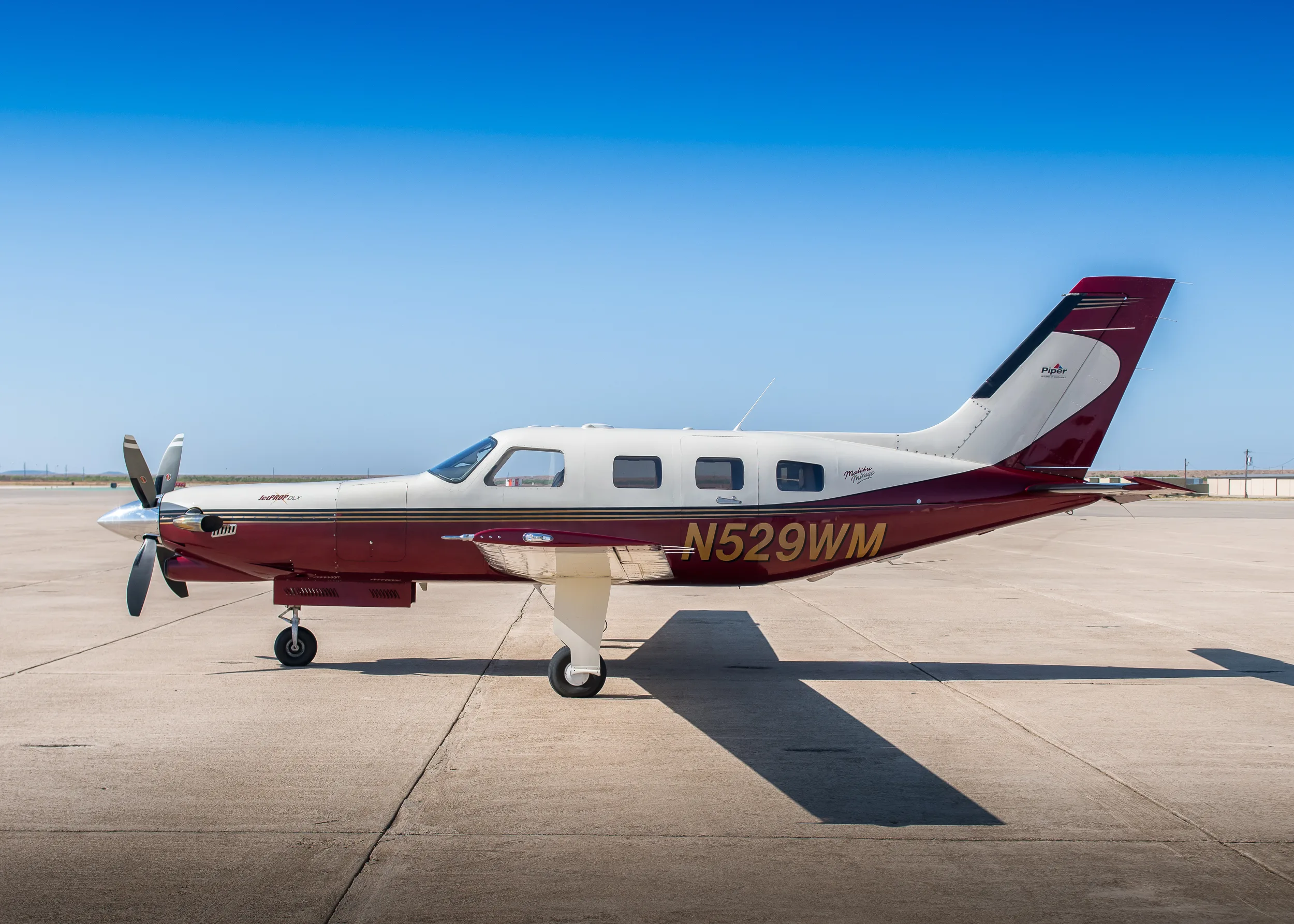 A side view of a maroon and white Piper M350 aircraft (N529WM) parked on an asphalt ramp under a clear blue sky.
