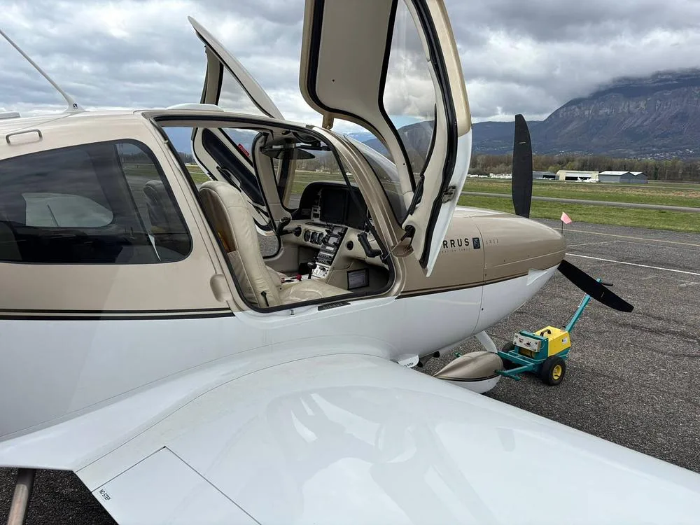 A Cirrus SR22 G3 aircraft with its gull-wing doors open is parked on an airfield tarmac with mountains in the background.