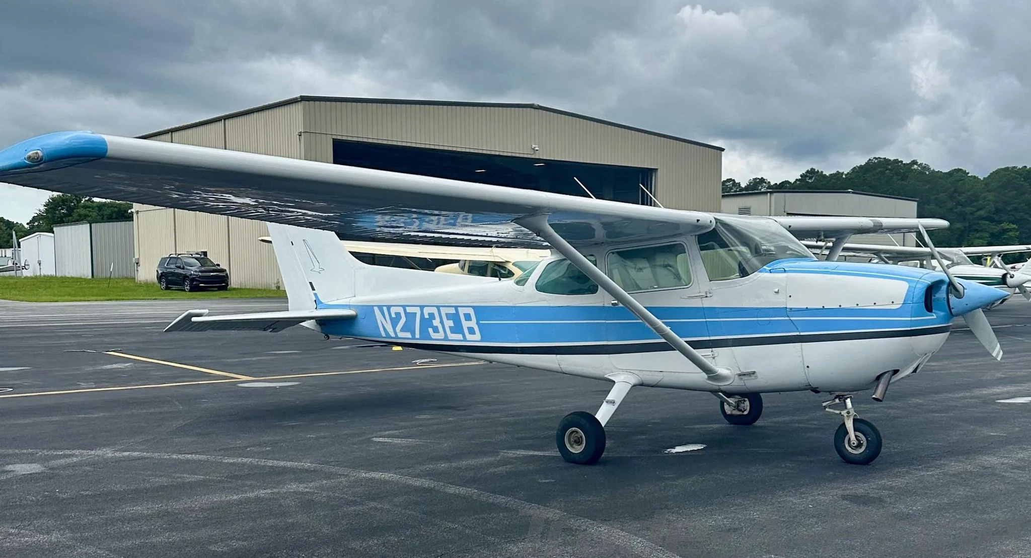 A white and blue Cessna 172, registration N273EB, parked on the tarmac with a hangar in the background.