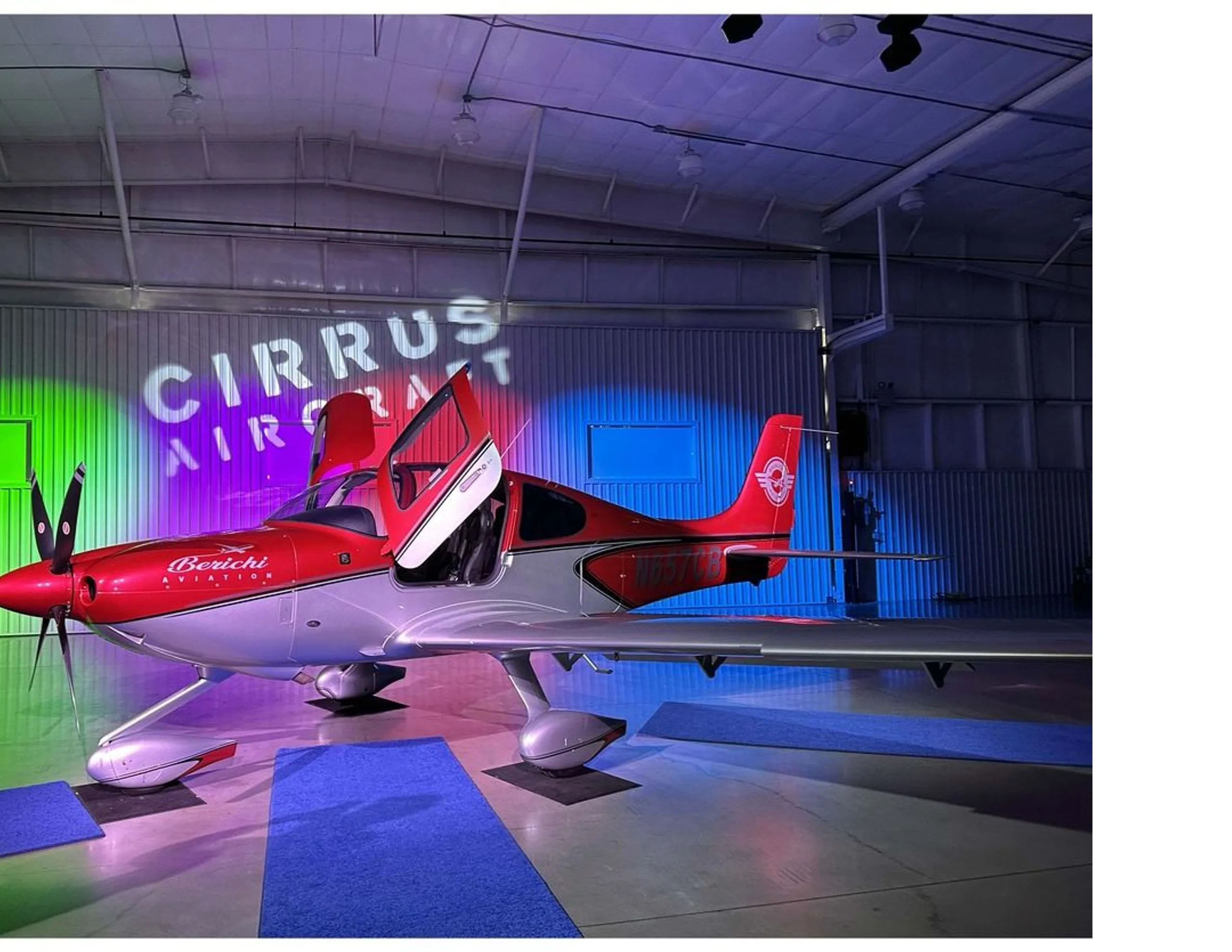 A side view of the red and silver Cirrus SR22 aircraft, N657CB, with open doors, in a hangar with "Cirrus Aircraft" projected on the wall.