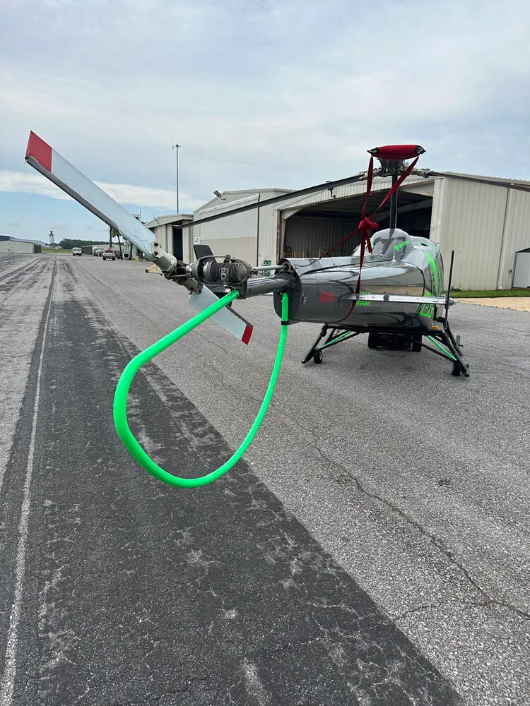 A front view of the gray Enstrom helicopter N88ZH with a bright green rotor blade tie-down on an airfield.