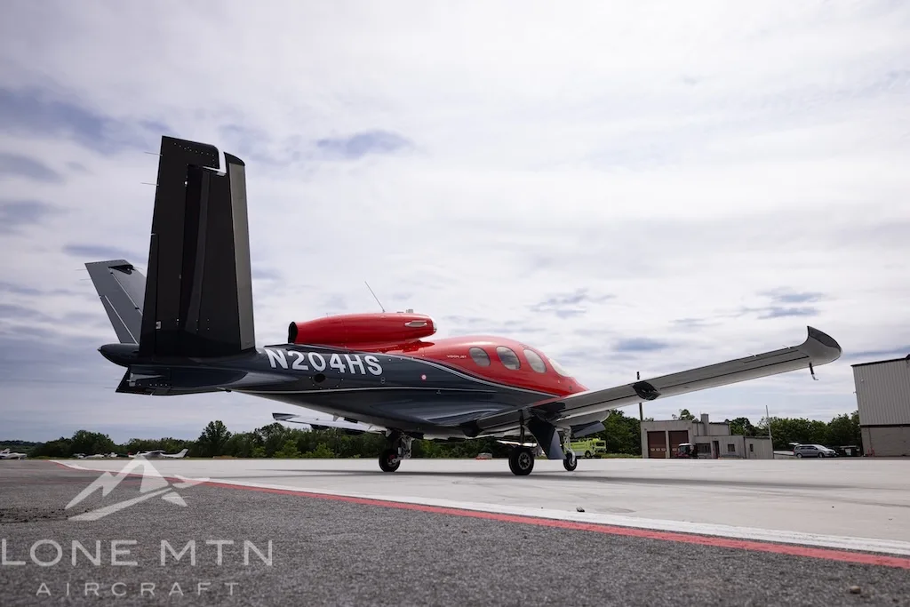 A rear-side view of the Cirrus Vision Jet N204HS in red and grey, with its distinctive V-tail, parked on an airfield.